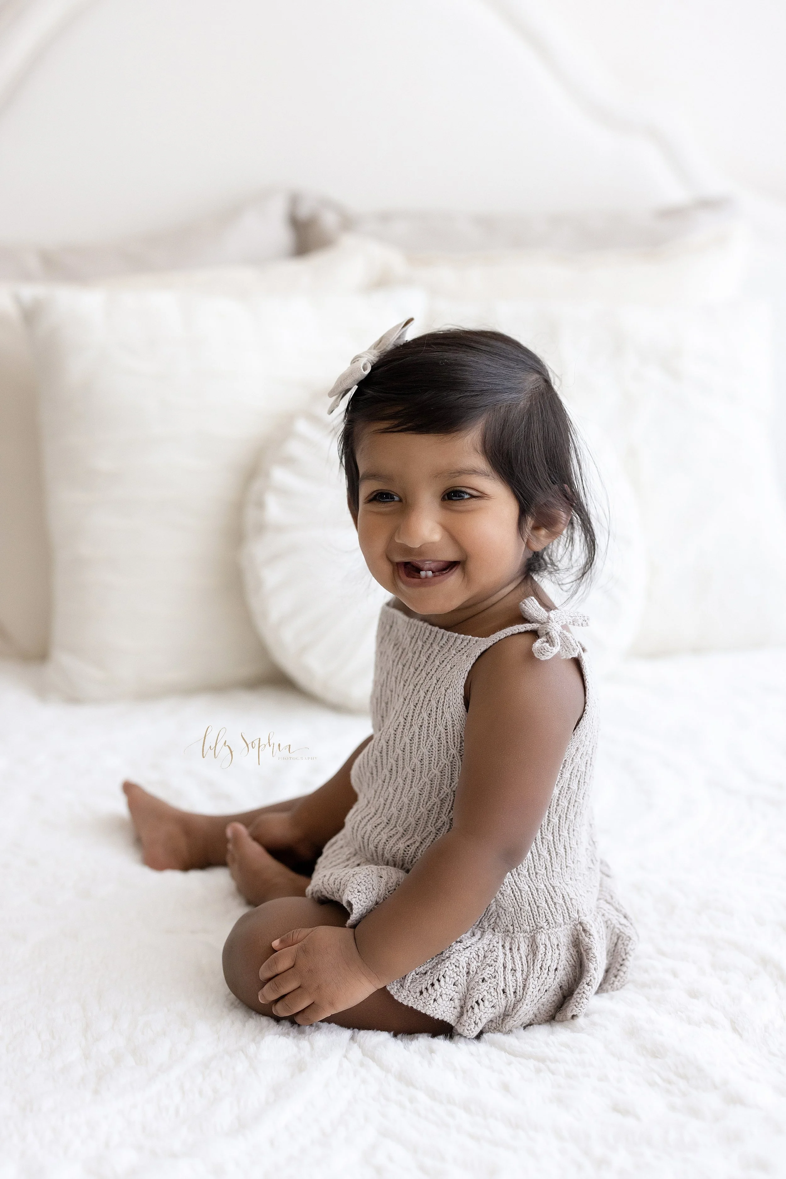 First birthday portrait of a one year old Indian girl who is smiling to show her two tiny bottom teeth while she sits on top of a bed in a natural light studio near Sandy Springs in Atlanta, Georgia. 