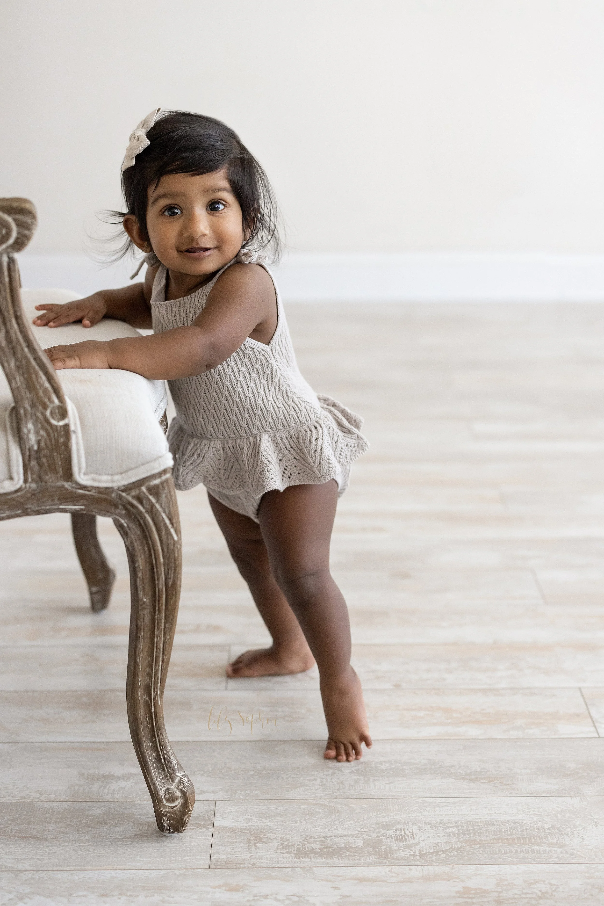  Milestone photo of a one year old Indian girl as she stands supporting herself on an upholstered armchair in a natural light studio near Buckhead in Atlanta. 