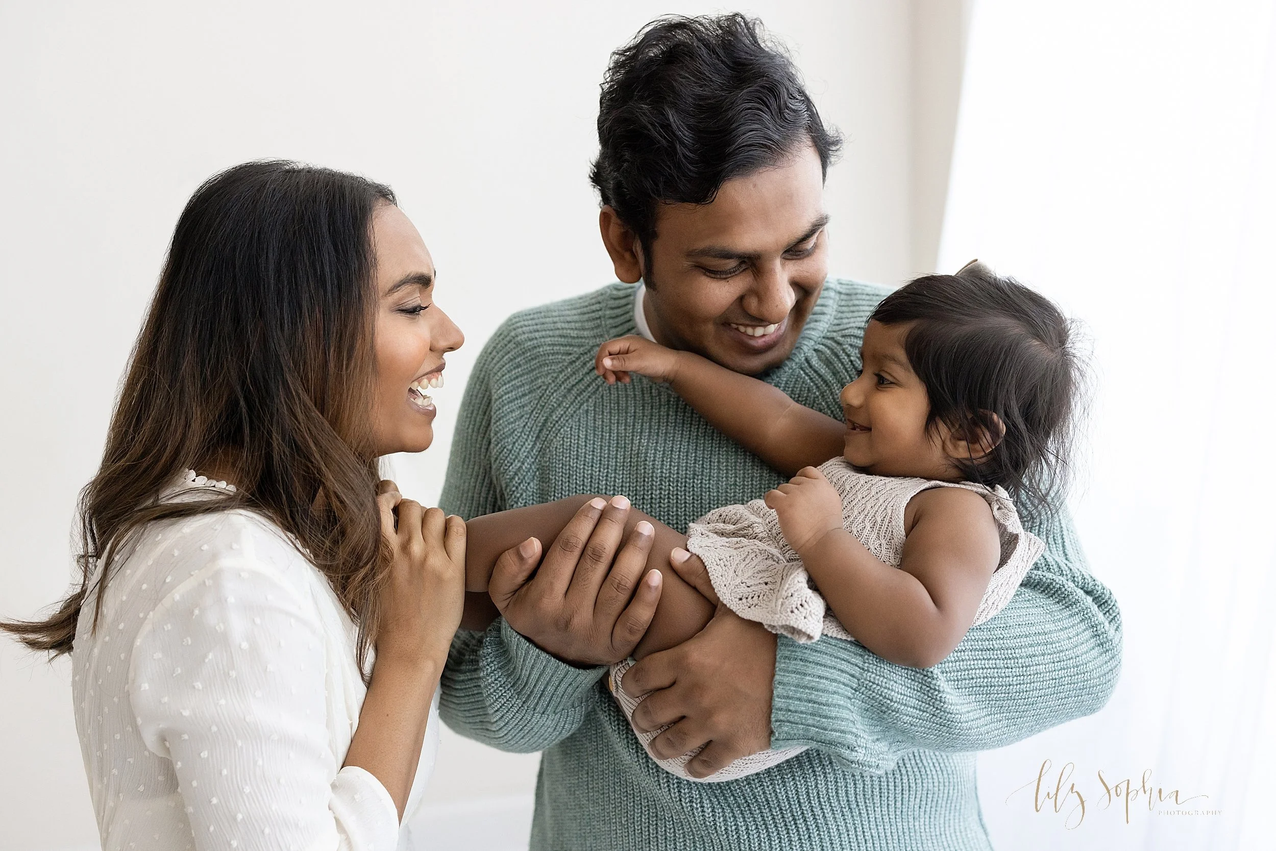  Family photograph of an Indian father holding his one year old daughter in his arms as mom faces her and holds her feet and the three of them interact with one another taken in a studio in front of a window streaming natural light near Morningside i
