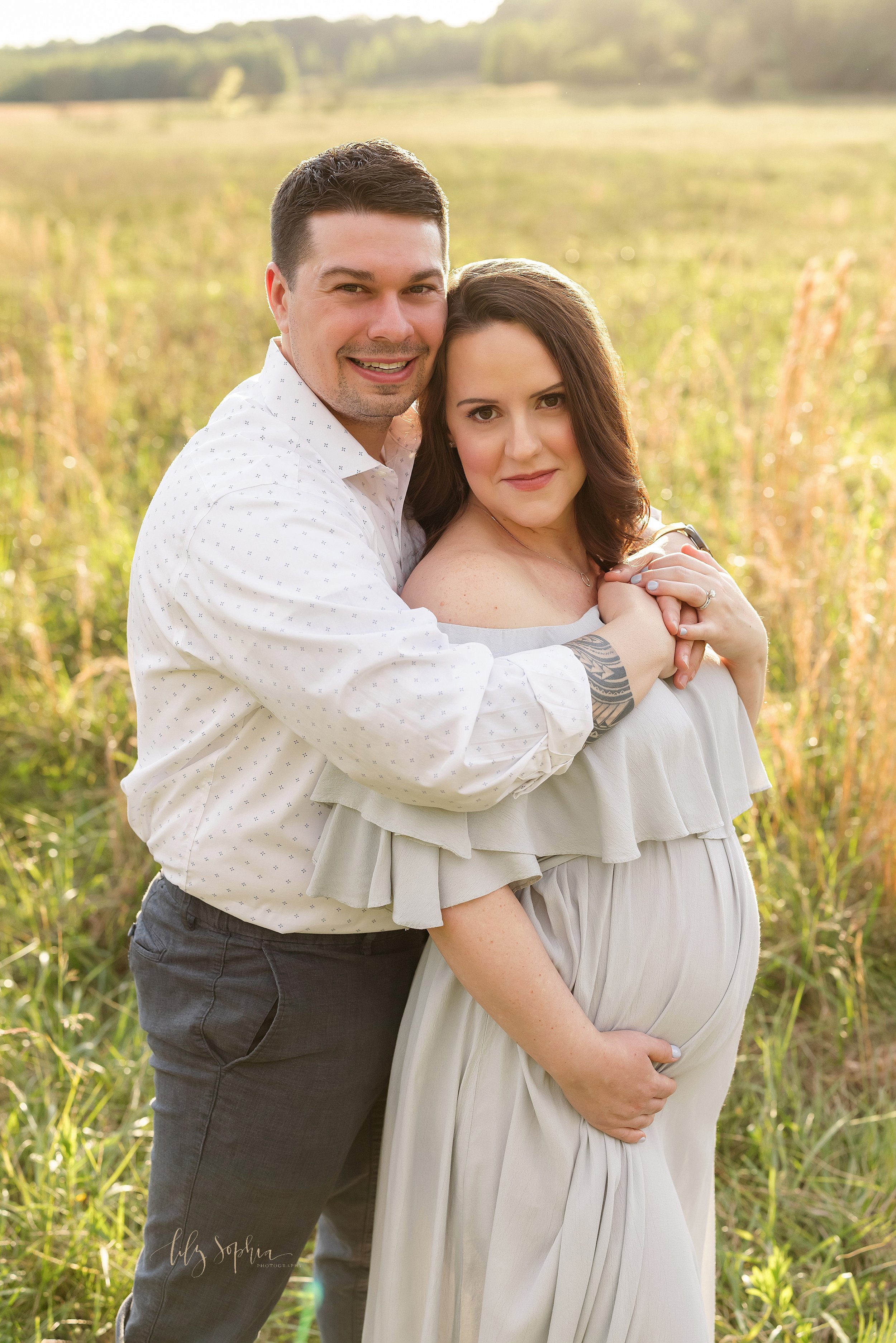  Maternity photo of a husband standing behind his wife and wrapping his arms around her as she holds her left hand on his and her right hand frames her belly as the couple stand at sunset in an Atlanta field. 