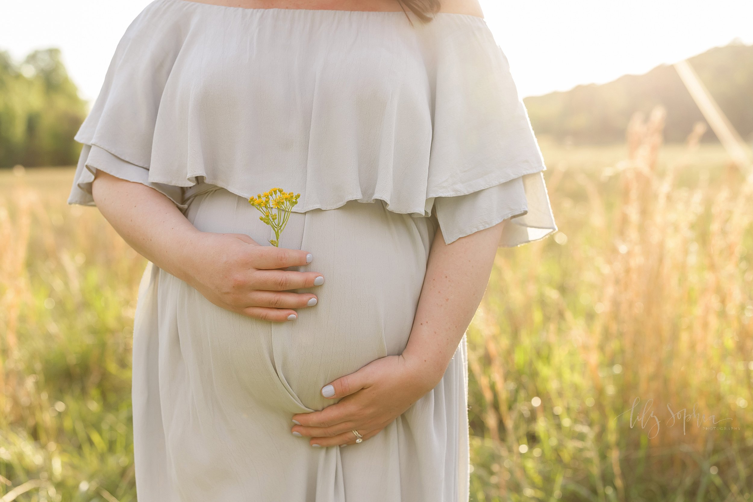  Maternity photo of a mother holding the base of her belly with her left hand as she holds a flowering piece of goldenrod in her right while stand in a field near Atlanta at sunset. 