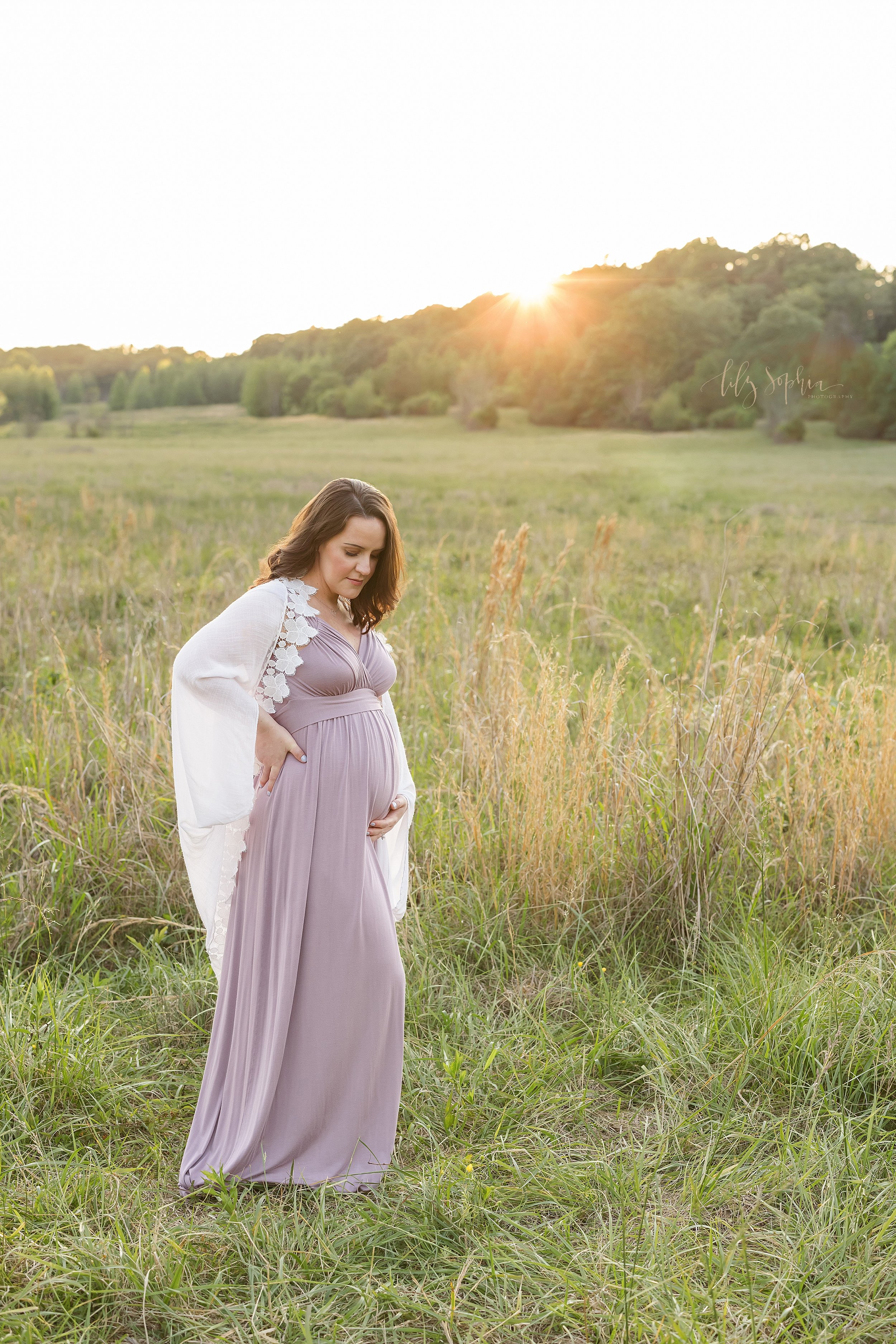  Maternity photo session of a pregnant mother wearing a jersey knit empire waist full-length gown and a white shawl draped over her shoulders as she holds her right hand on her hip and her left hand holds the base of her belly while she stands at sun