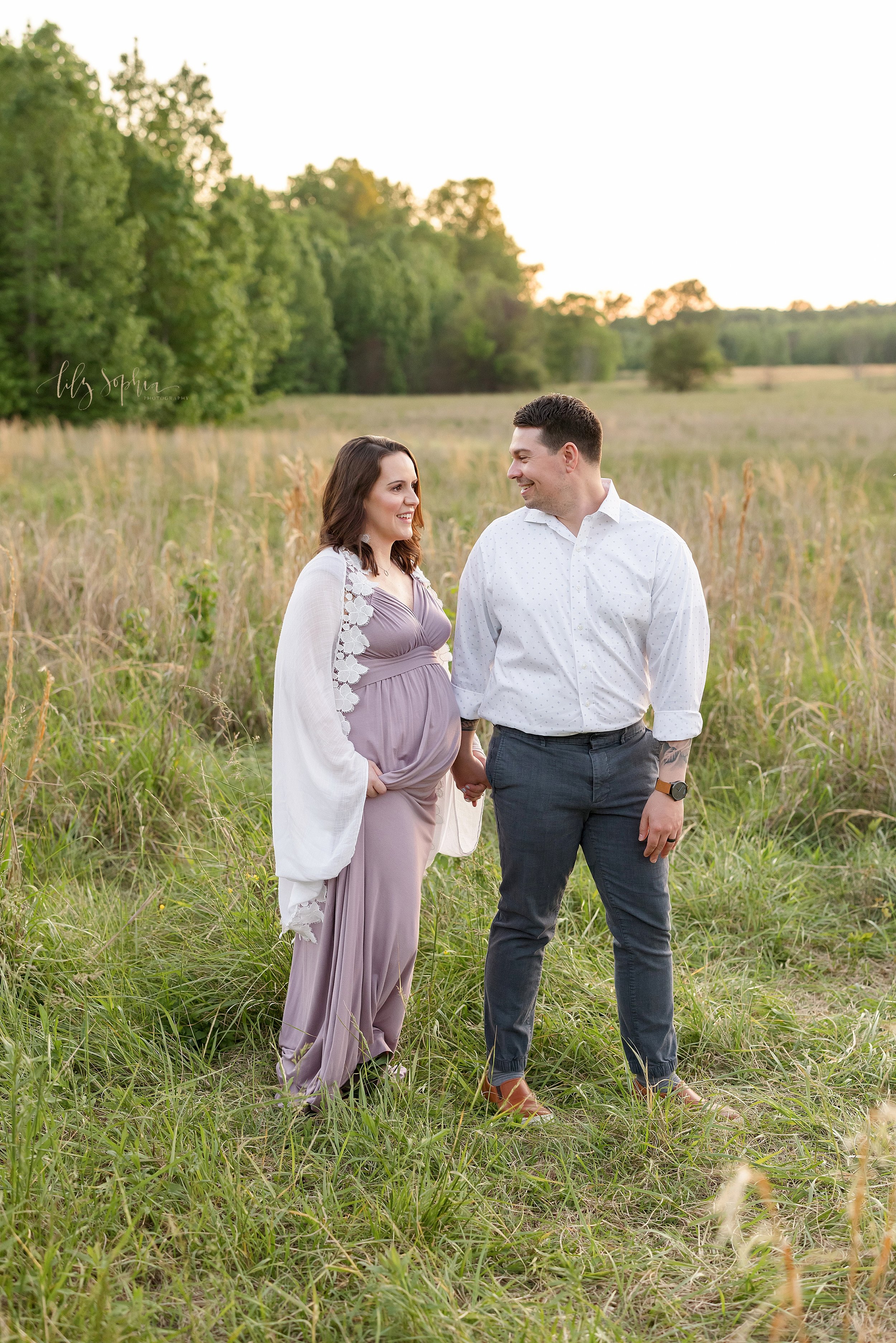  Maternity photo of a husband holding his pregnant wife’s hand as he leads her through a field near Atlanta at sunset. 