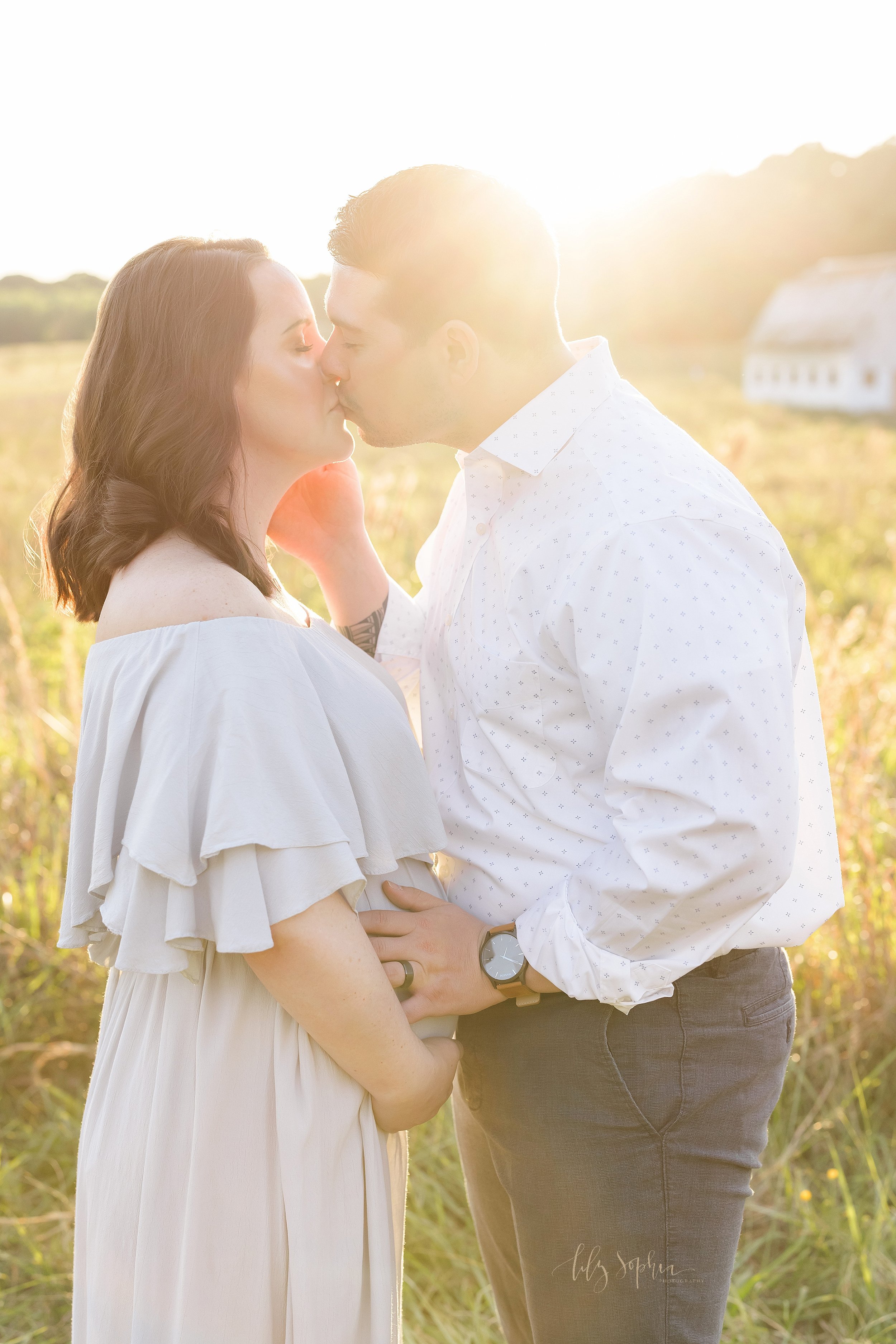  Maternity photograph of a pregnant mother standing facing her husband in an Atlanta field at sunset as she holds her belly with her hands and her husband kisses her while holding his hand on his child in utero. 