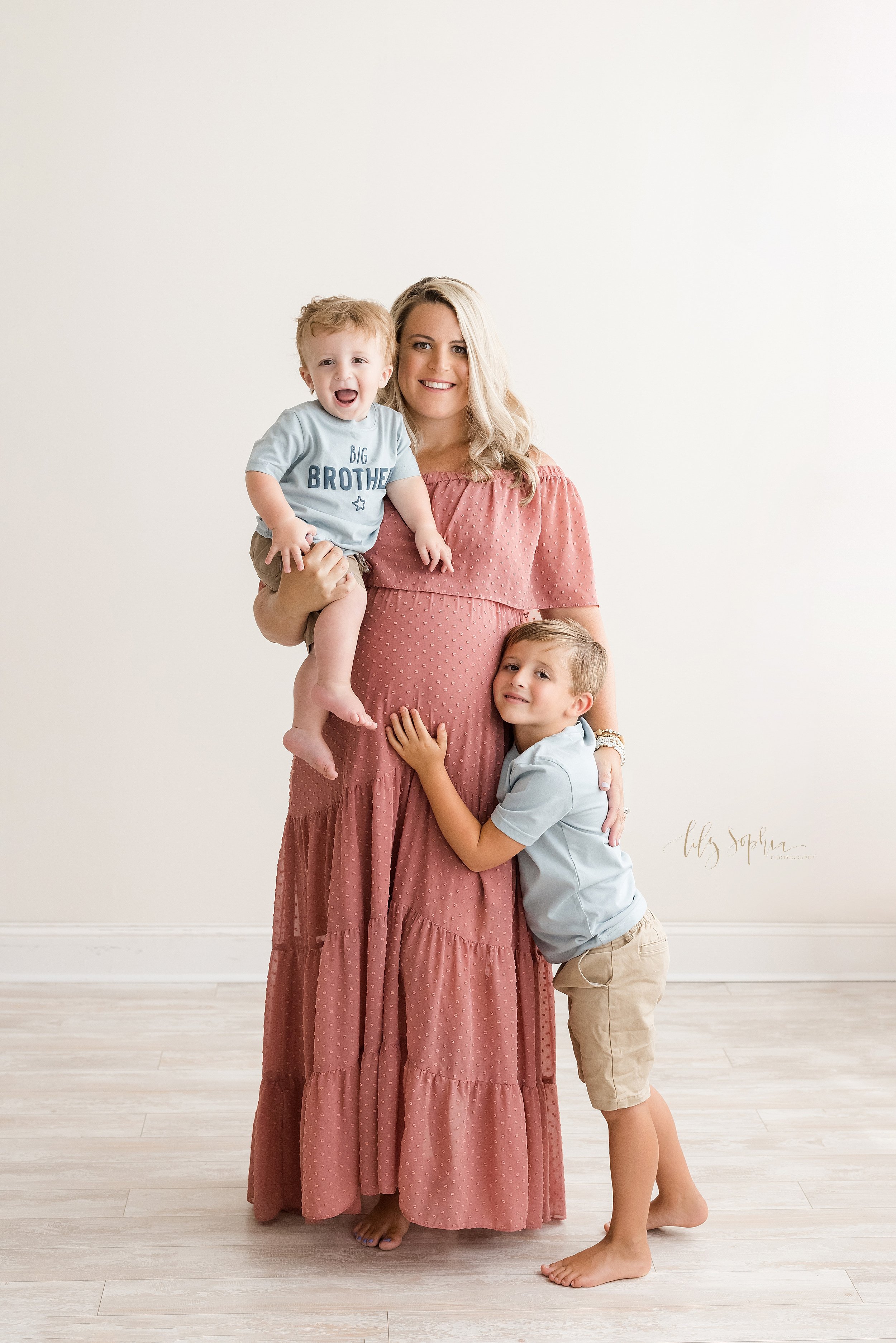  Maternity photo session with mom holding her one year old son in her arms as her other young son stands by her side and touches his sibling in utero taken in a studio using natural light near Kirkwood in Atlanta. 