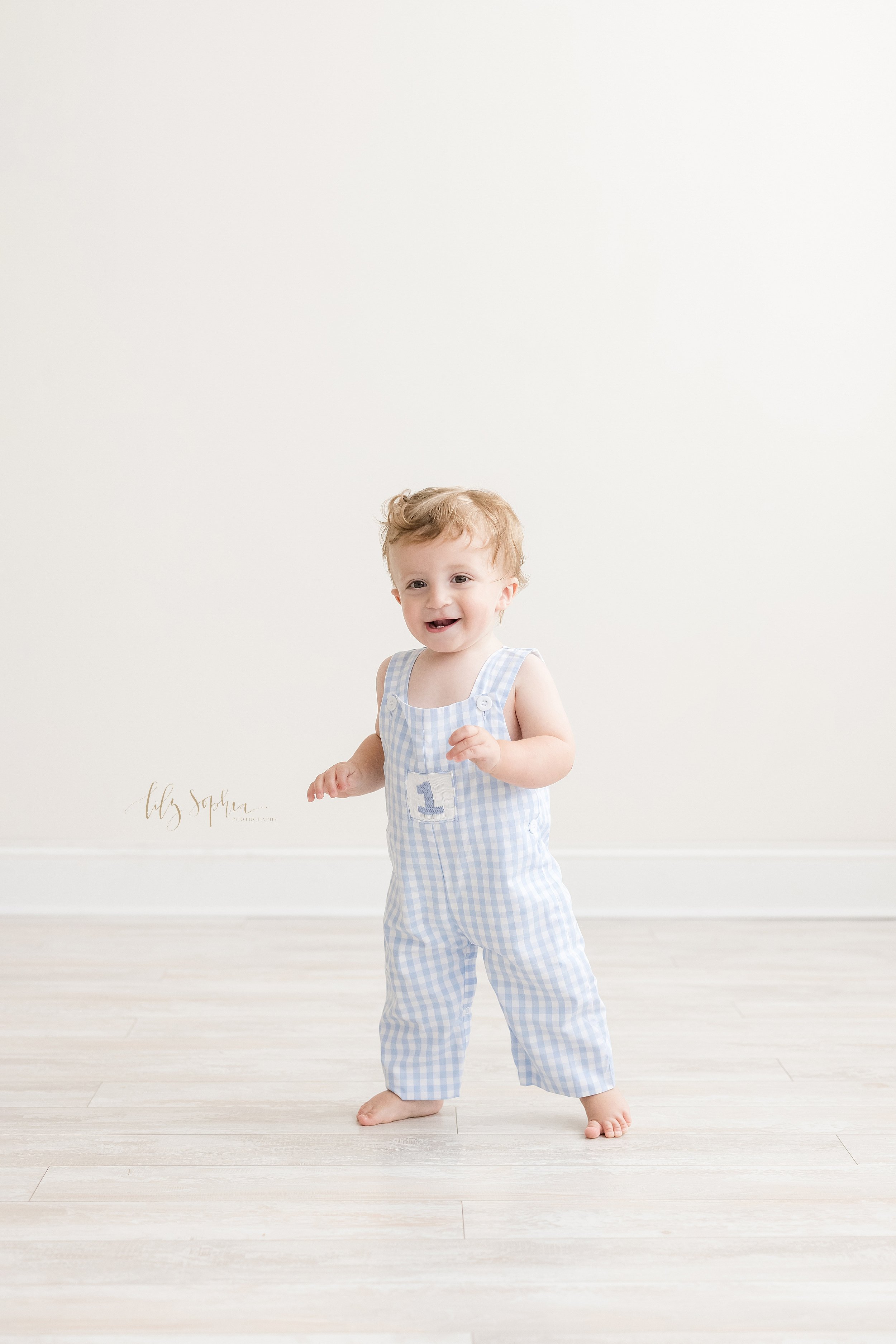  Milestone photograph of a one year old little boy as he toddles in blue and white gingham overalls in a natural light studio near Morningside in Atlanta. 