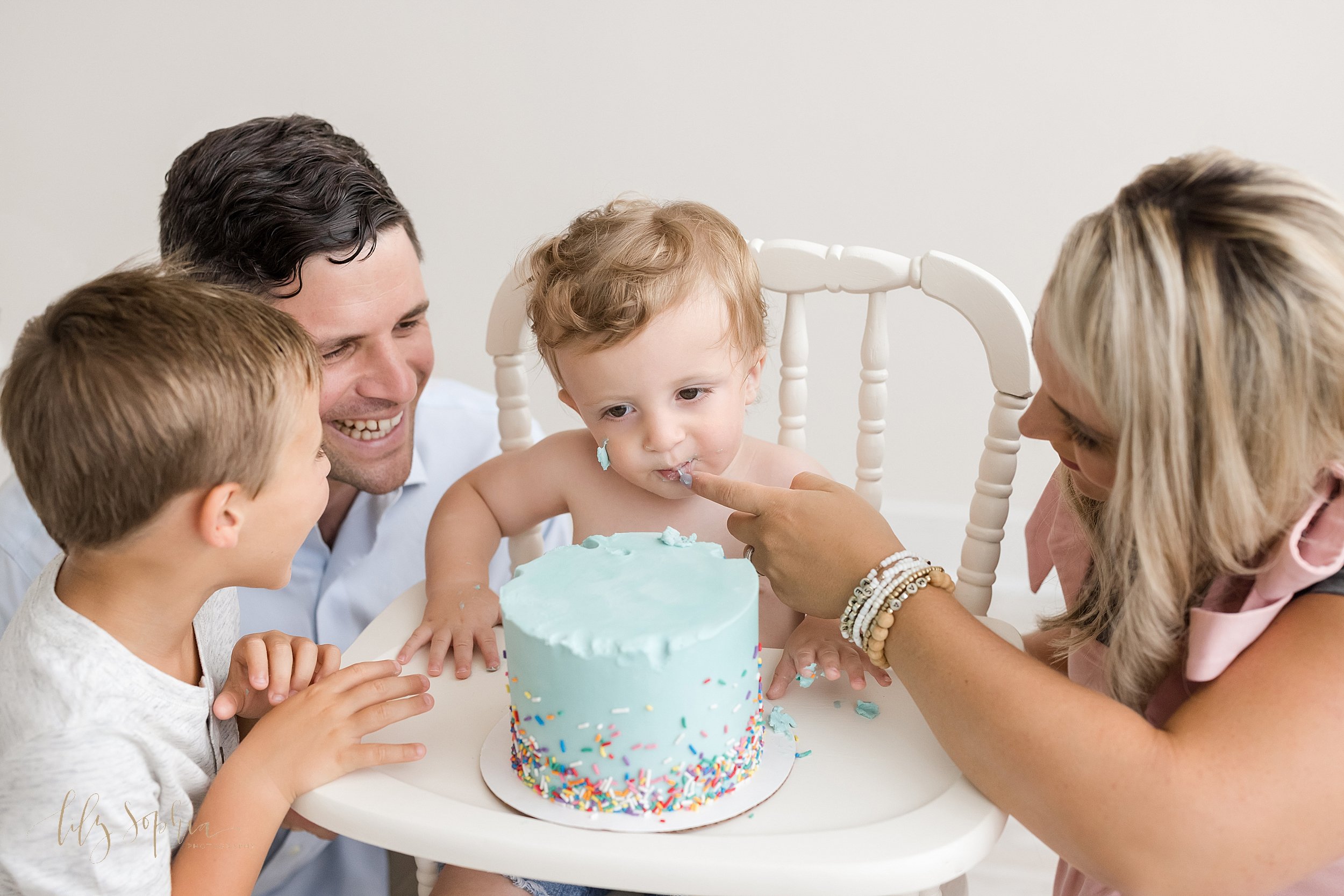  First birthday session with a one year old little boy and his family as he sits in a wooden antique highchair with his smash cake on the tray as he mother squats to his left and feeds him a taste of icing and his father and brother watch on as dad s