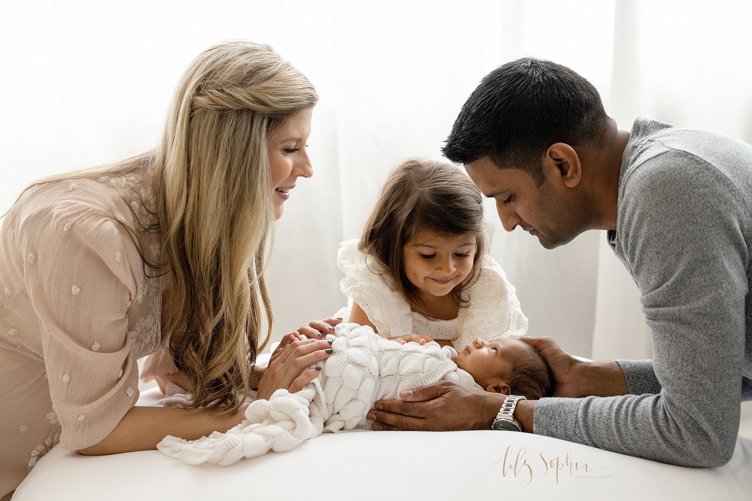  Family newborn portrait of an awake newborn lying on his back on a bed while mom, dad, and sister lean over to see him taken in front of a window streaming natural light in a studio near Poncey Highlands in Atlanta. 