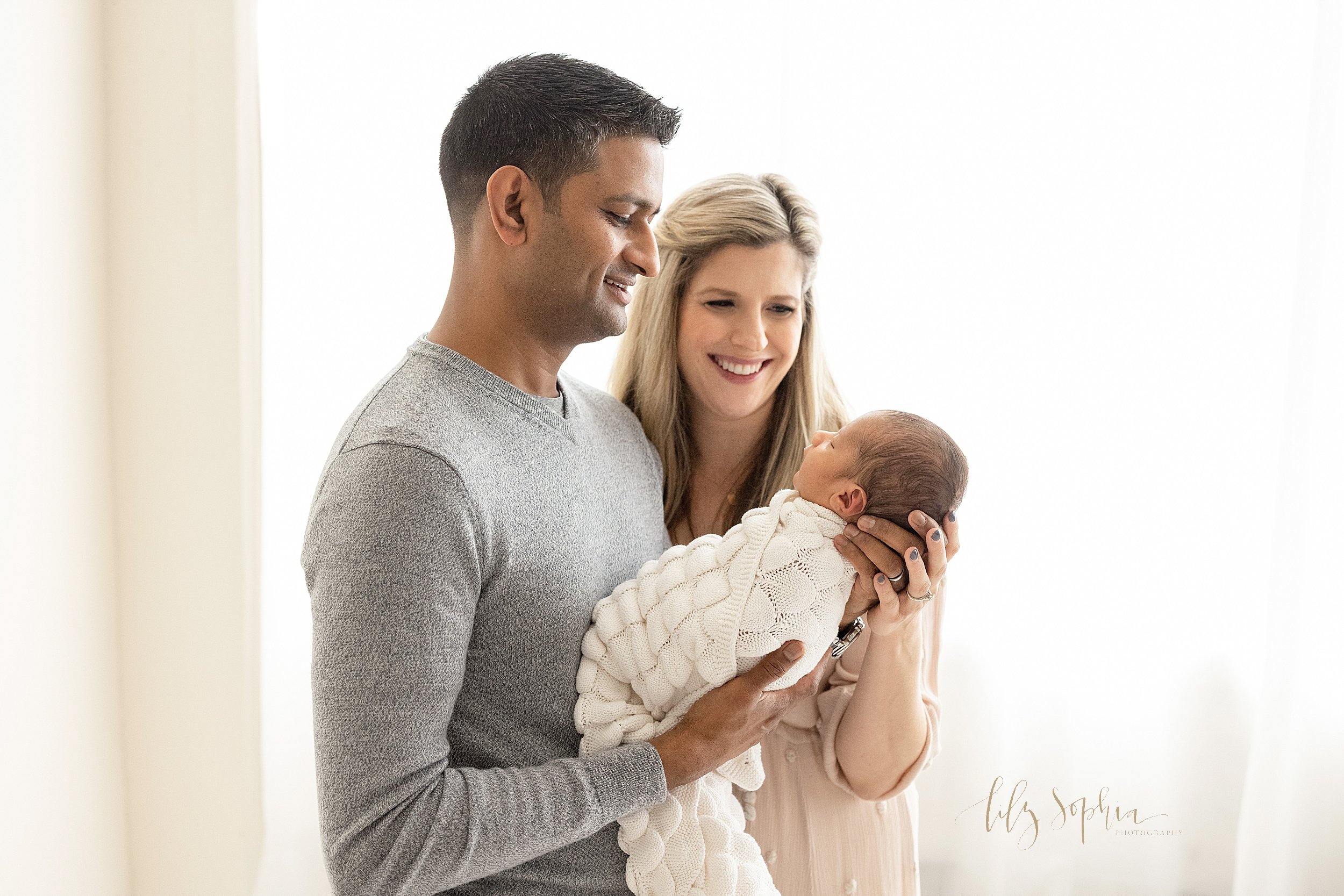  Newborn family photograph of a father holding his infant son in his hands as his wife stands to his left and the two of them smile proudly taken in  front of a window streaming natural light in a studio near Vinings in Atlanta. 
