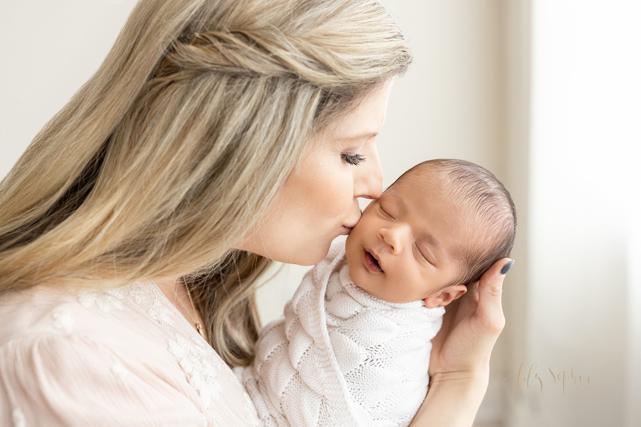  Newborn photograph of a mother holding her newborn baby son in her hand and kissing his cheek as he sleeps swaddled to his chin in a white crocheted blanket taken in a studio near Buckhead in Atlanta that uses natural lighting. 