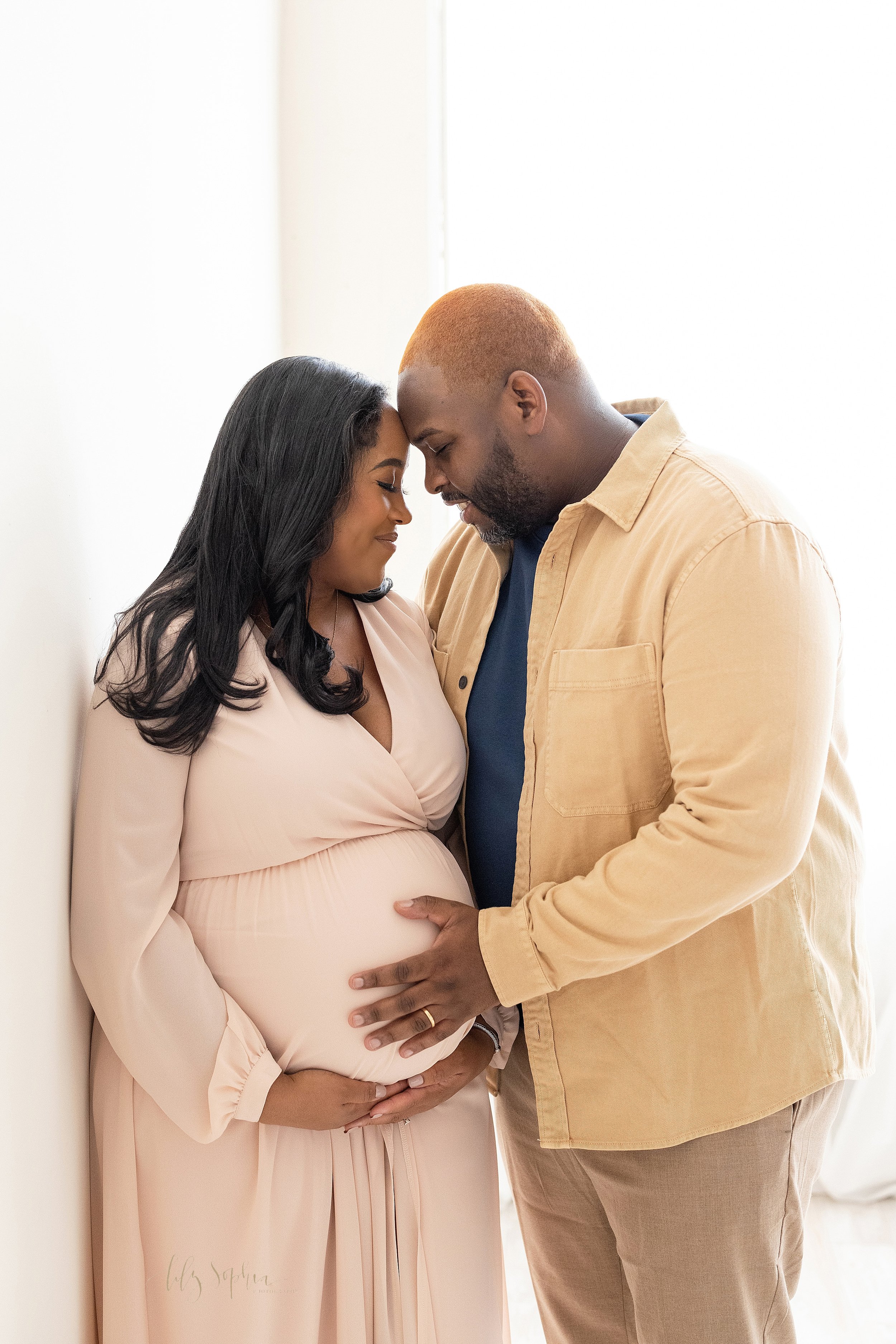  Maternity photo session in a natural light studio of a pregnant mother and her husband as they stand with their foreheads together while she frames her belly with her hands and he places his hand on their child in utero taken near Oakhurst in Atlant