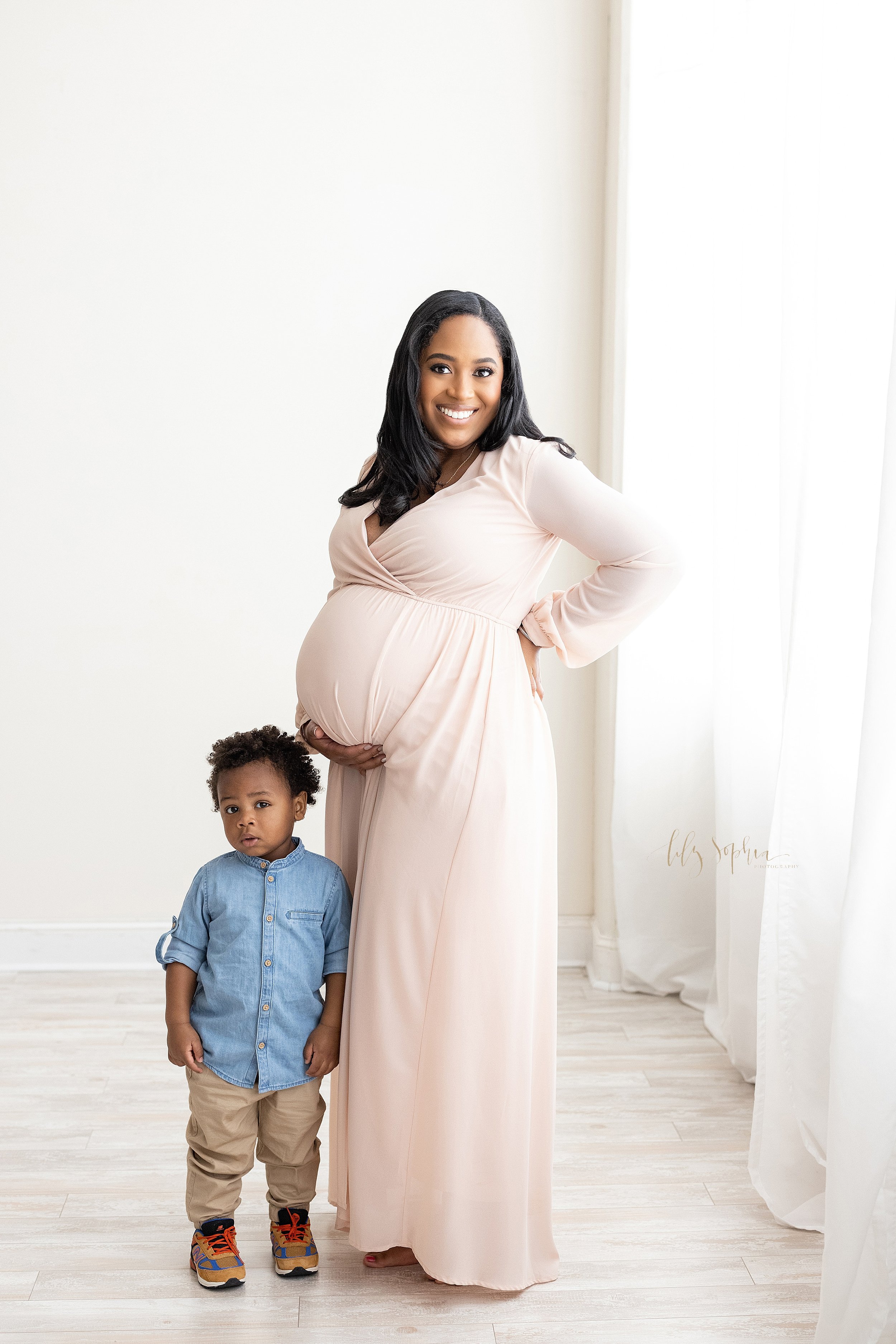  Maternity photo session in a natural light studio of a pregnant African-American mother standing and holding the base of her belly with her right hand and holding her hip with her left hand as her toddler son stands in front of her taken near Old Fo
