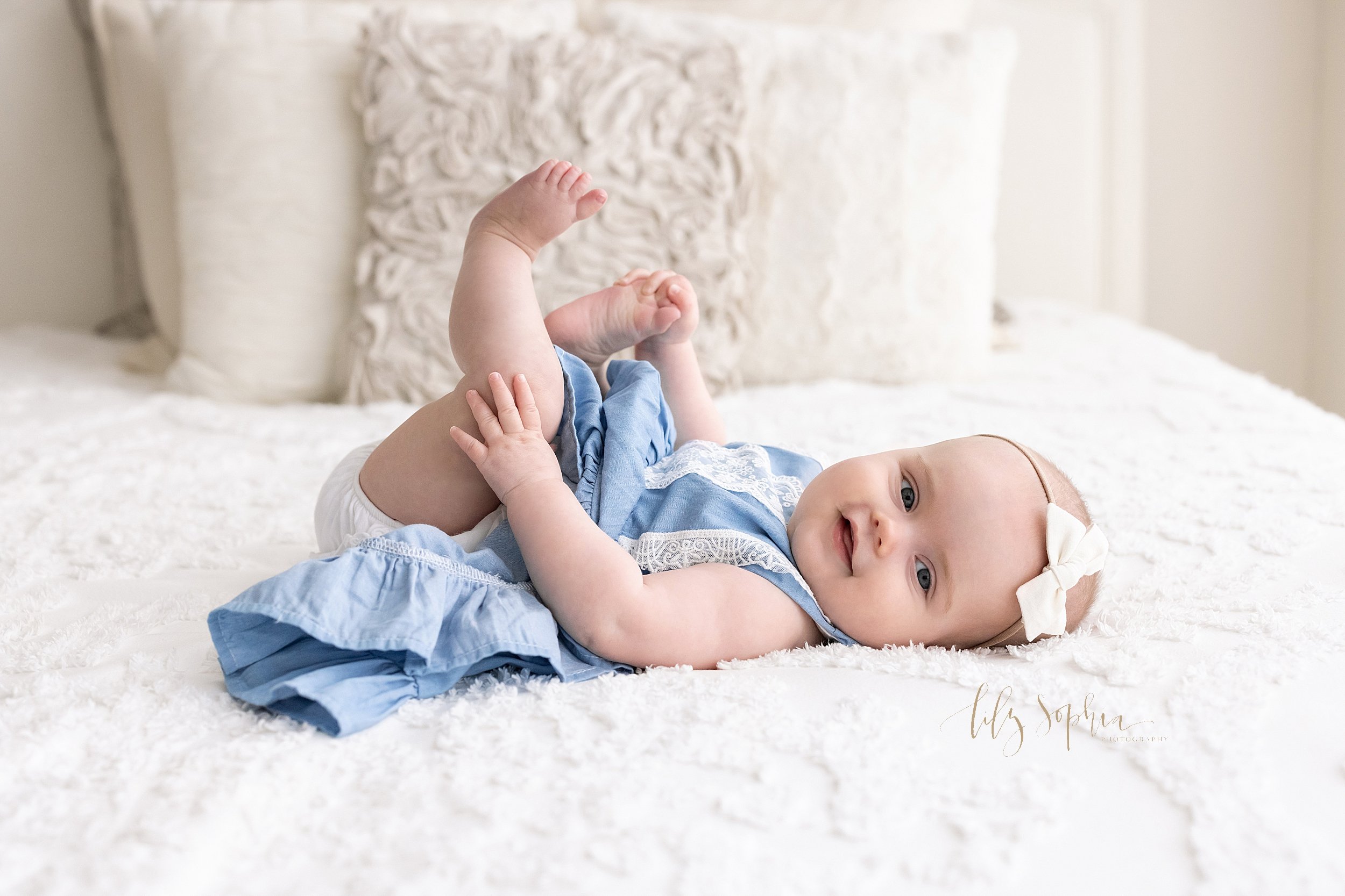  Milestone photo of a six month old little girl lying on her back on a bed in a natural light studio holding her right foot with her right hand and placing her left hand on her left knee taken near Midtown in Atlanta. 
