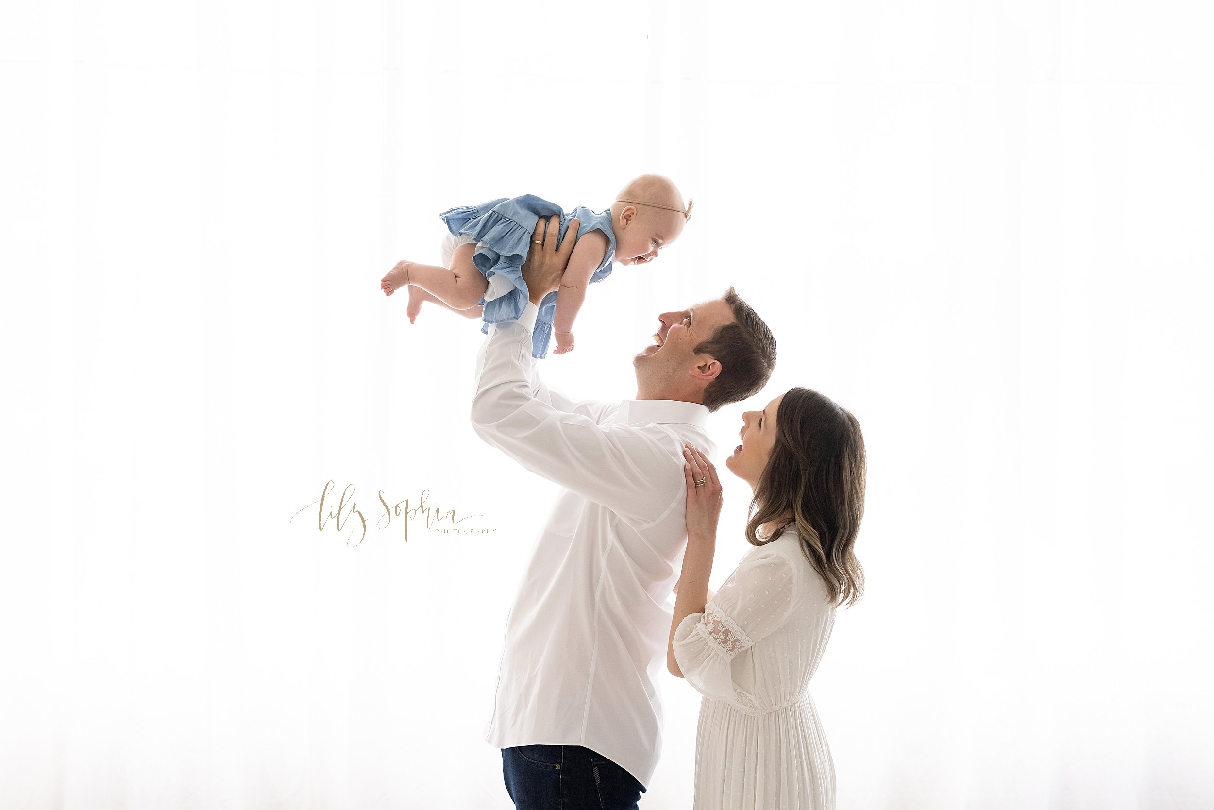  Family photo of a father holding his six month old baby daughter above his head as she laughs and mom stands behind her husband with her right hand on his shoulder while they smile at their daughter as they stand in front of a window streaming natur