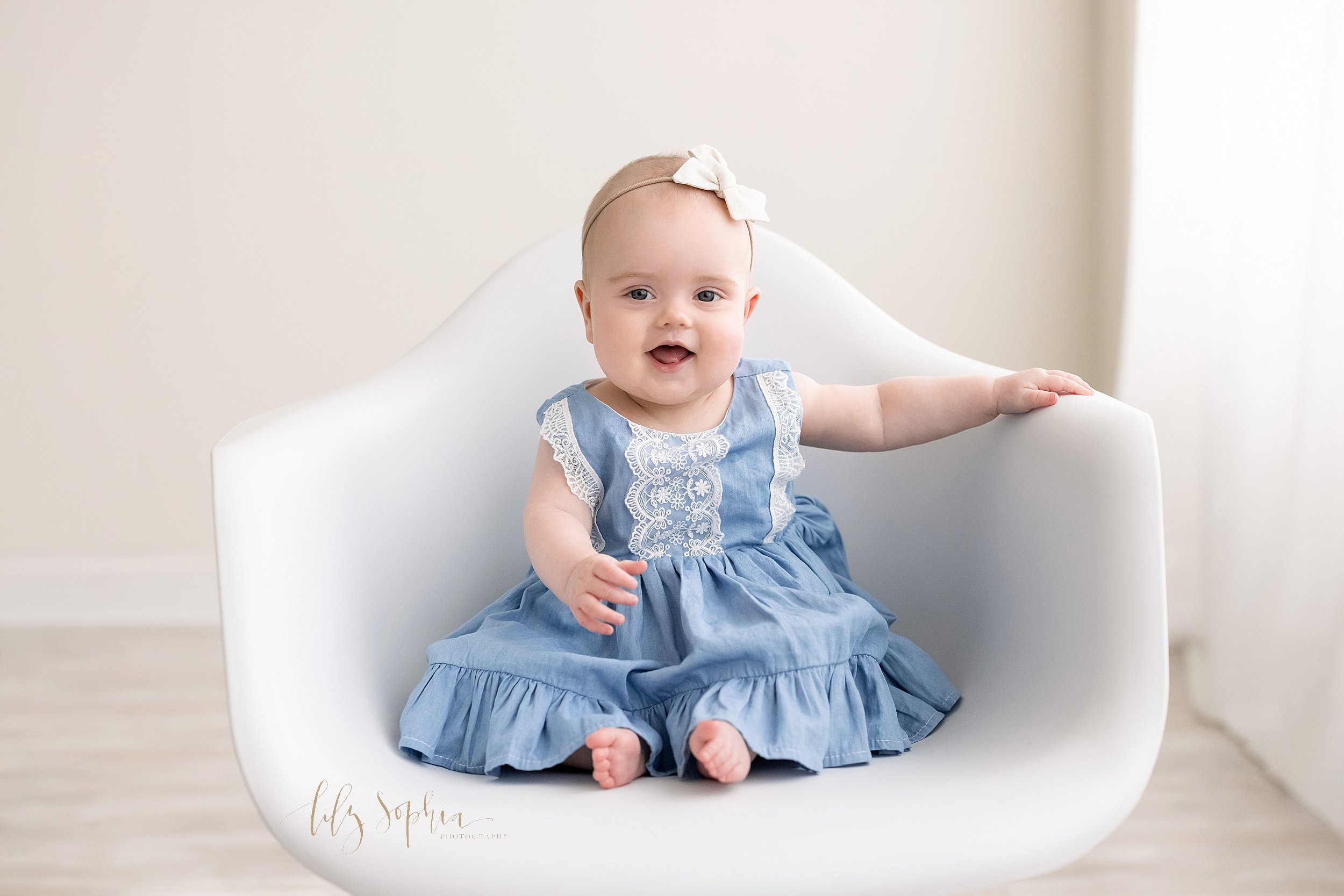  Milestone baby portrait of a six month old baby girl as she sits in a molded white chair smiling toothless while she holds onto the chair with her left hand taken in a natural light studio near Alpharetta in Atlanta, Georgia. 