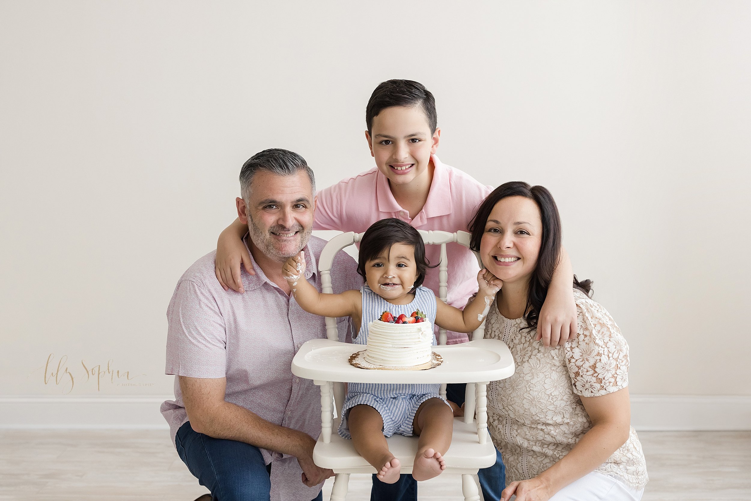  Family portrait of a family celebrating their son's first birthday as the one year old sits in a wooden antique high chair with a smash cake on the tray and his parents squat on either side of him as he puts his icing covered hands under their chins