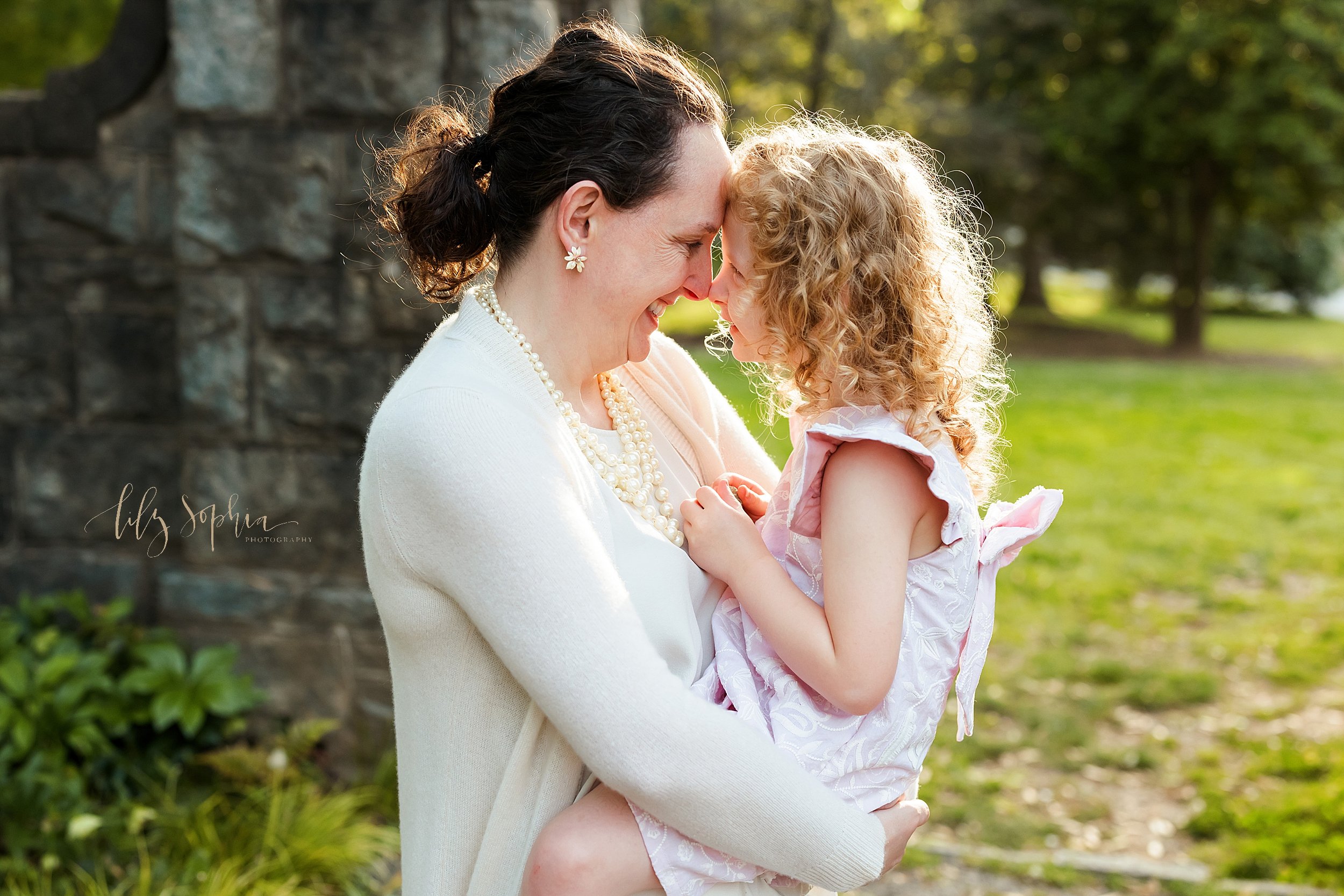  Family portrait of a mother holding her young daughter in her arms as the two of them put their foreheads together and talk while mom stands in a stone entranceway in a park at sunset near Atlanta, Georgia. 