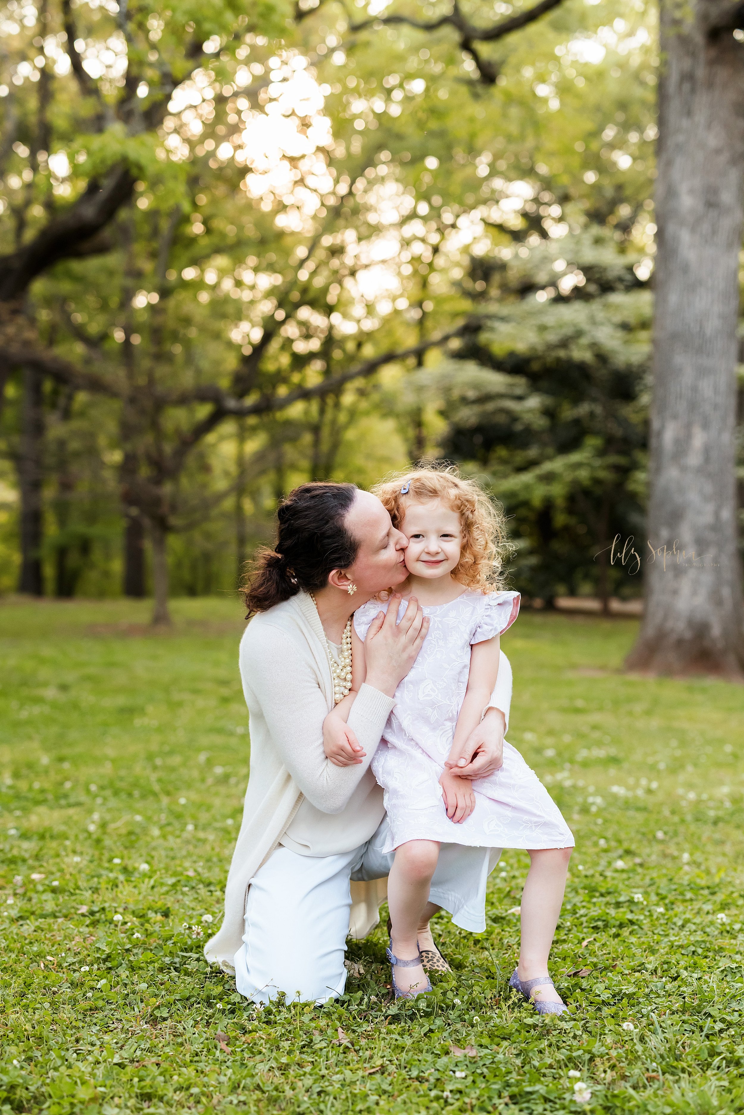  Family photo shoot of a mother with her curly red-headed daughter as mom kisses her on the cheek while she sits on her mom’s knee taken in a park near Atlanta, Georgia at sunset. 