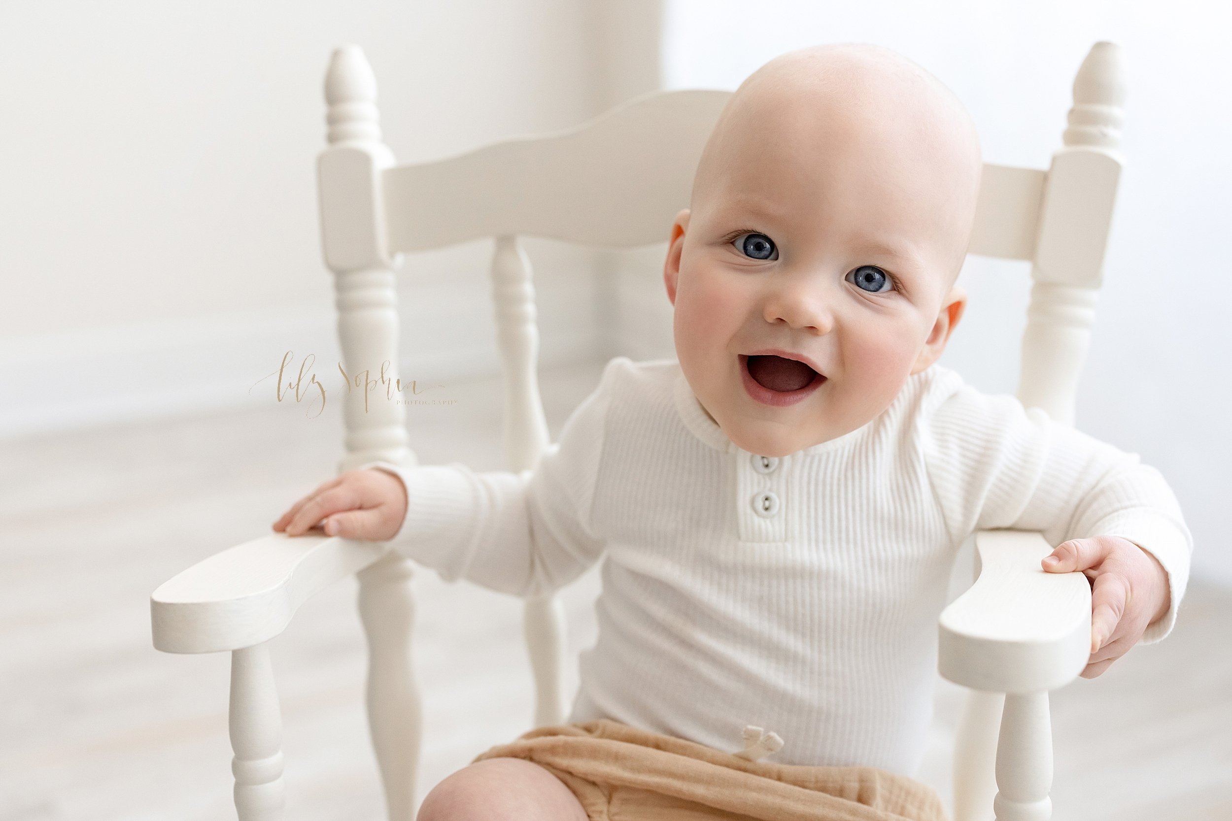  Milestone baby portrait of a happy eight month old boy as he proudly sits in a white rocking chair in front of window streaming natural light in a studio near Alpharetta in Atlanta, Georgia. 