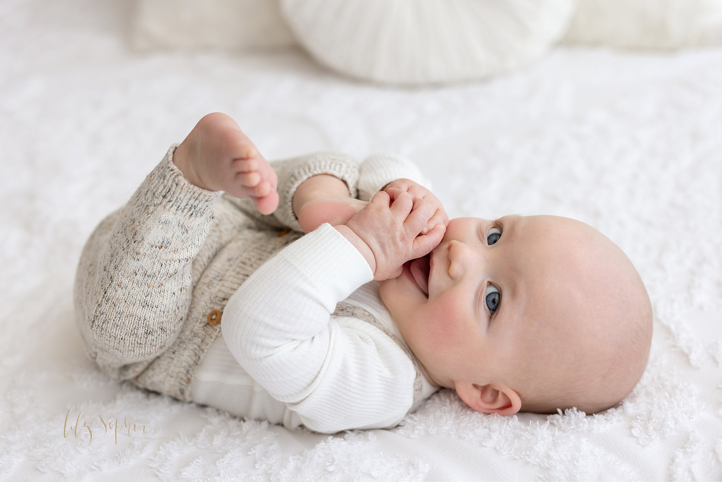  Milestone portrait of an eight month old little boy as he lies on his back on top of a bed chewing on the toes of his right foot taken in a natural light studio near Buckhead in Atlanta. 