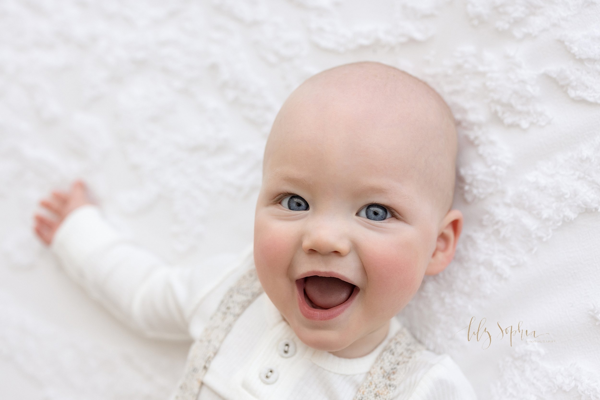 Close-up portrait of an excited blue-eyed eight month old baby boy as he laughs with his mouth open while lying on his back on a bed in a studio near Vinings in Atlanta, Georgia that utilizes natural light. 