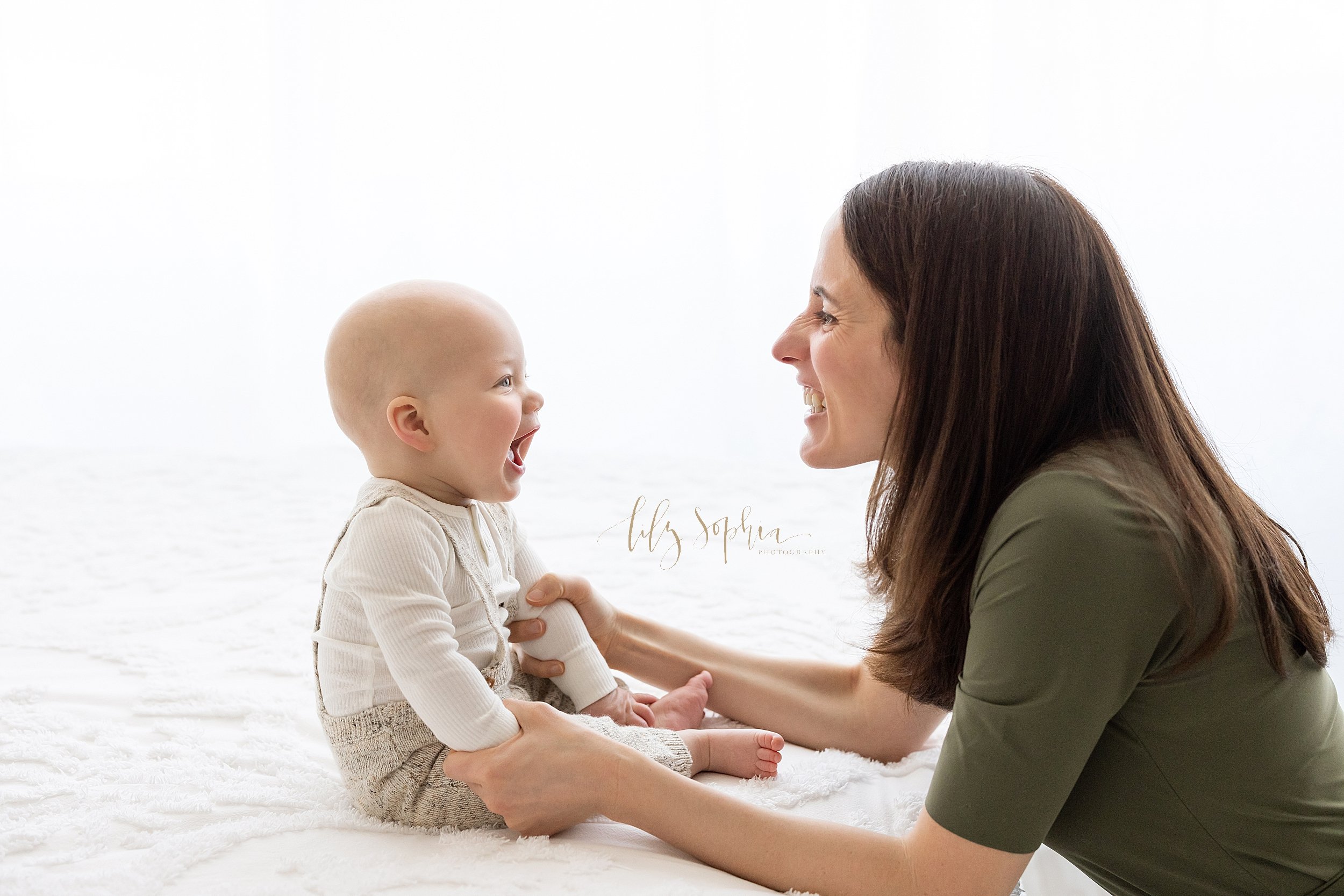  Family photograph of an eight month old little boy sitting at the edge of a bed with his mother facing him as the two of them laugh and talk taken in front of a window streaming natural light near Smyrna in Atlanta in Lily Sophia Photography Studios
