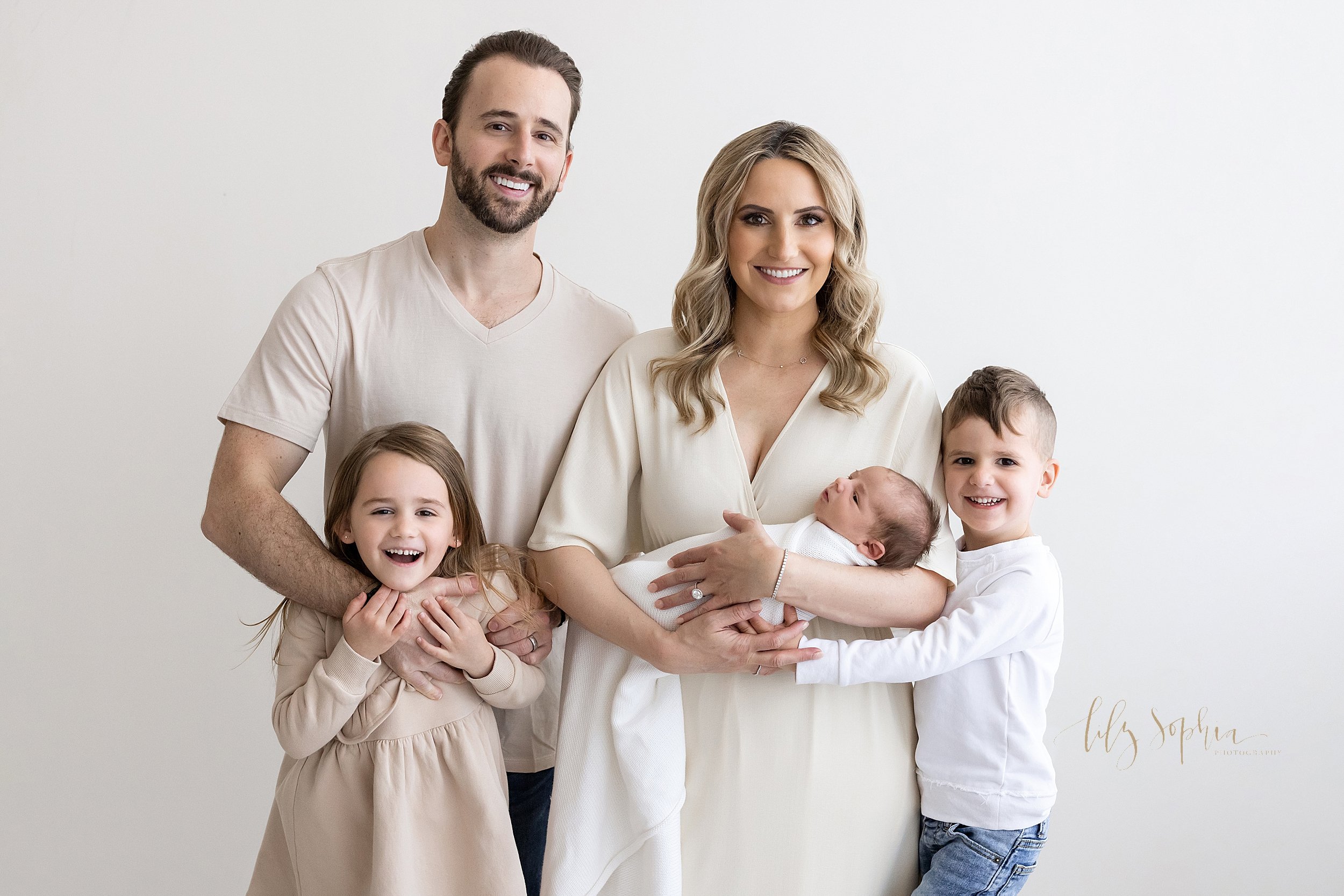  Family portrait of a mother holding her newborn son in her arms with her husband standing to her right  with their daughter standing in front of her father as he puts his right and around the chest of his daughter and their son stands to the left of