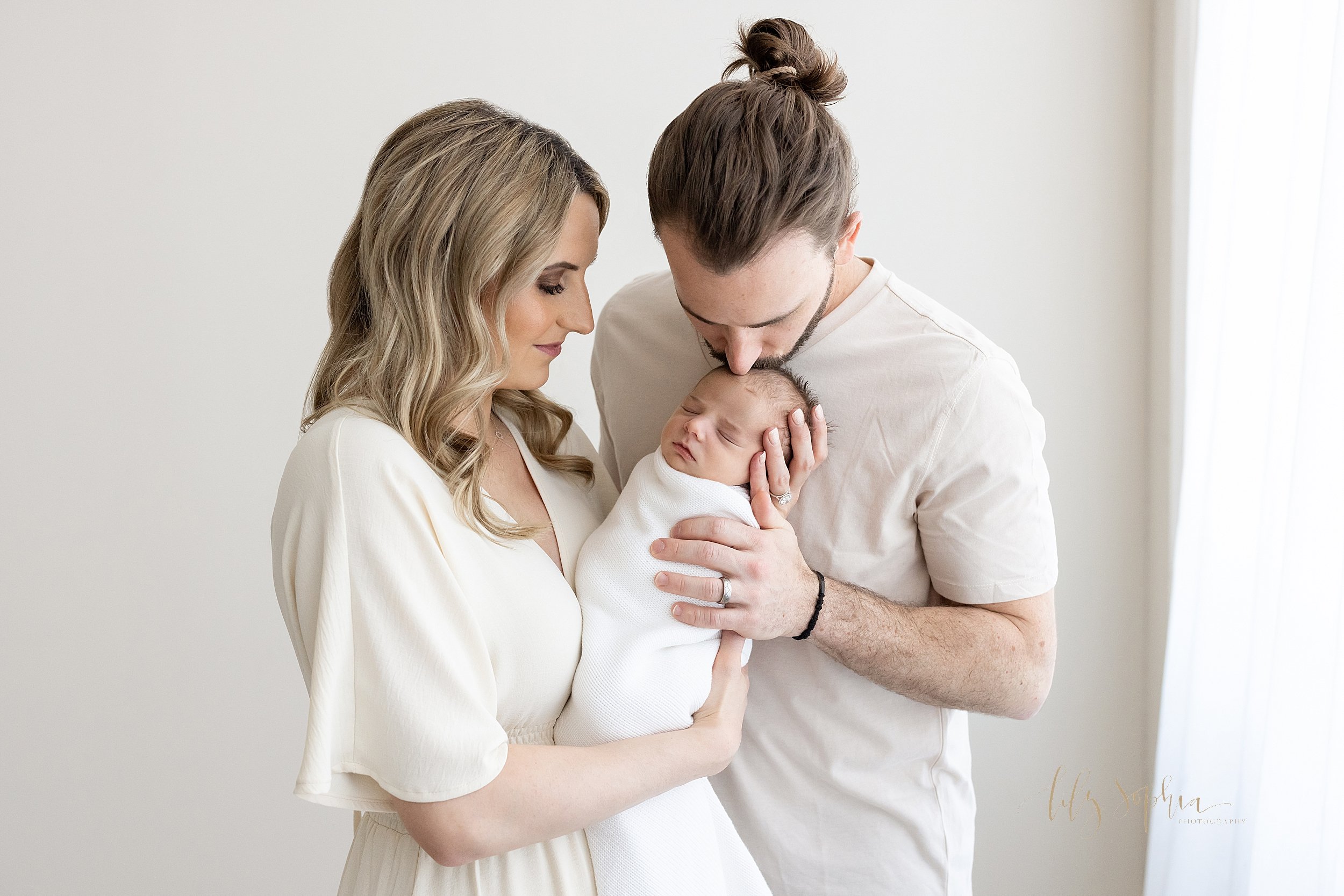  Family newborn photo of a mother holding her infant son in her hands as her husband stands next to her on her left side and kisses the crown of his son’s head taken in front of a window streaming natural light in a studio near Poncey Highlands in At