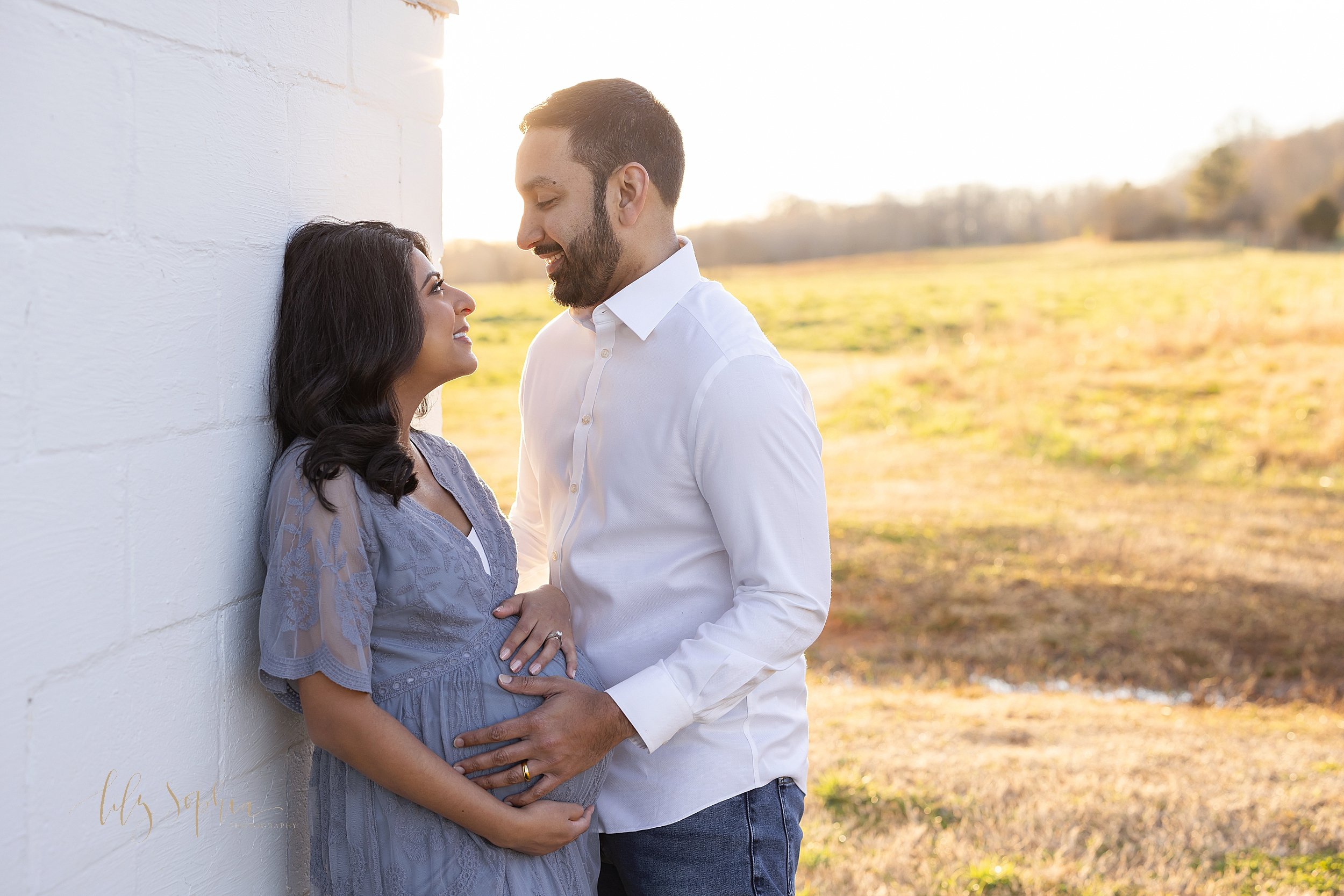  Maternity portrait of a pregnant mother standing with her back against a white concrete wall holding the base of her belly with her right hand and resting her left hand on top of her belly as her husband faces her and looks into her eyes while he pl