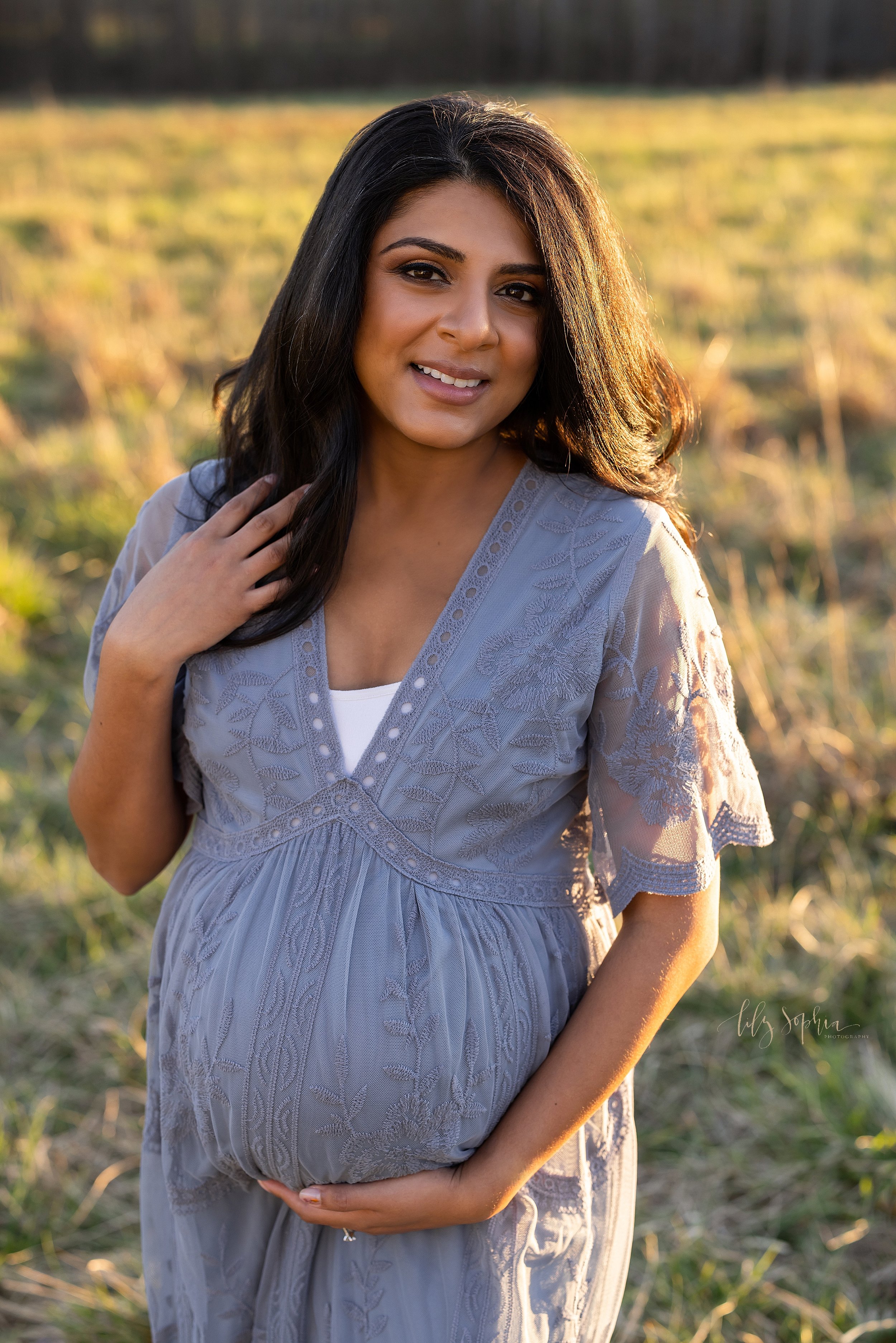  Maternity portrait of a mother wearing a V-necked blue embroidered gown with short bell sleeves as she is standing in an Atlanta field with her right hand holding the base of her belly and her left hand touching her hair as the sun sets. 