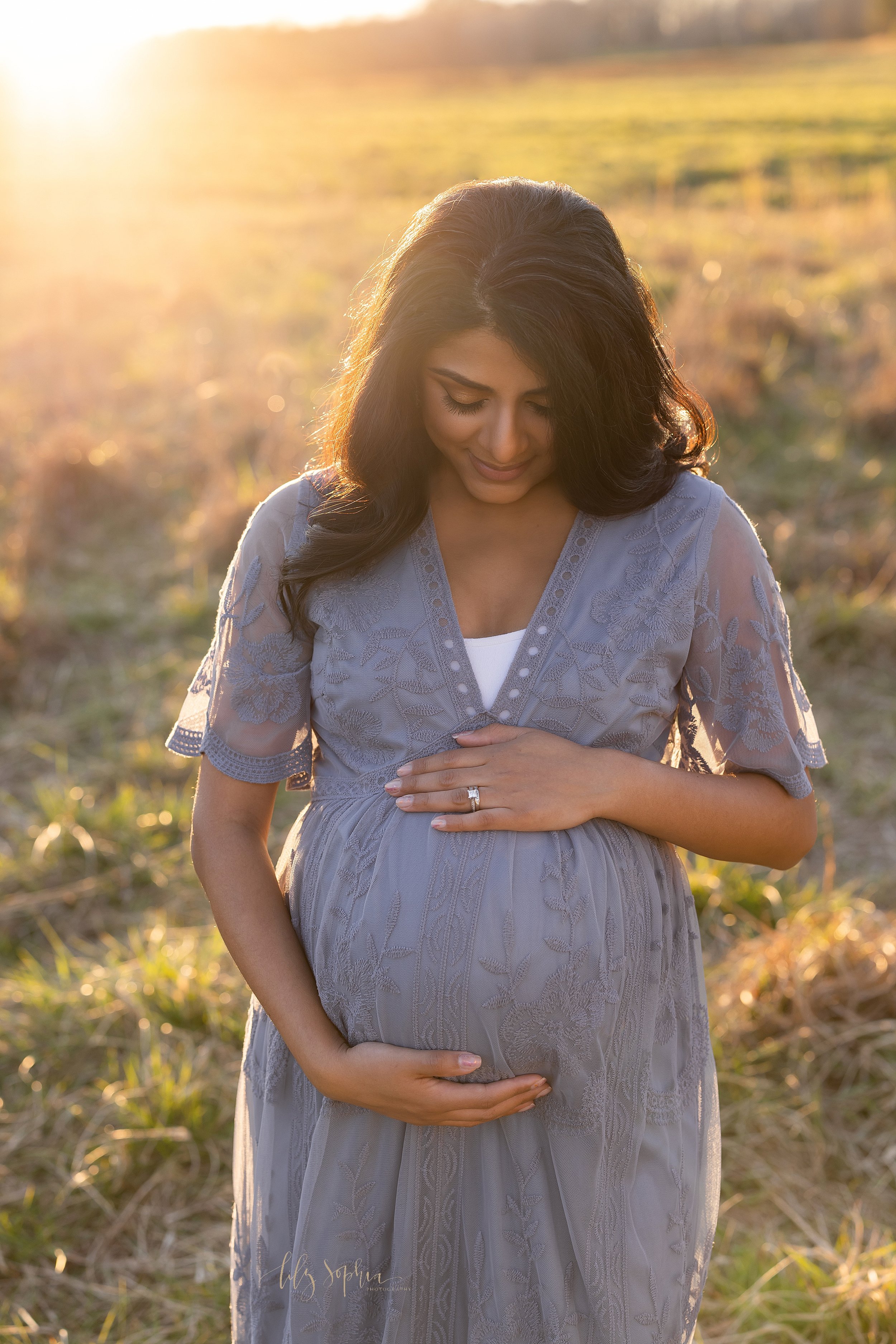  Maternity picture of a pregnant mother standing in a field as the sun sets behind her during autumn with her hands framing her belly while she looks down at her baby in utero and smiles taken near Atlanta. 