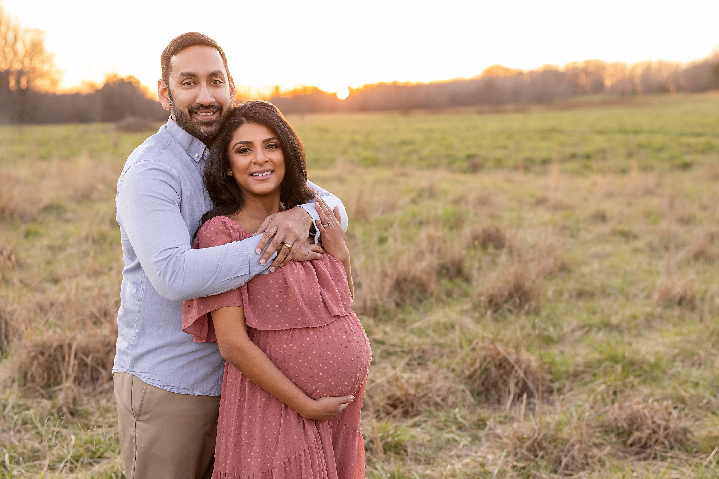  Maternity photo session of a husband and wife as they stand in a field near Atlanta with the husband standing behind his wife wrapping his arms around her chest and the wife holding the base of her belly with her right hand and placing her left hand