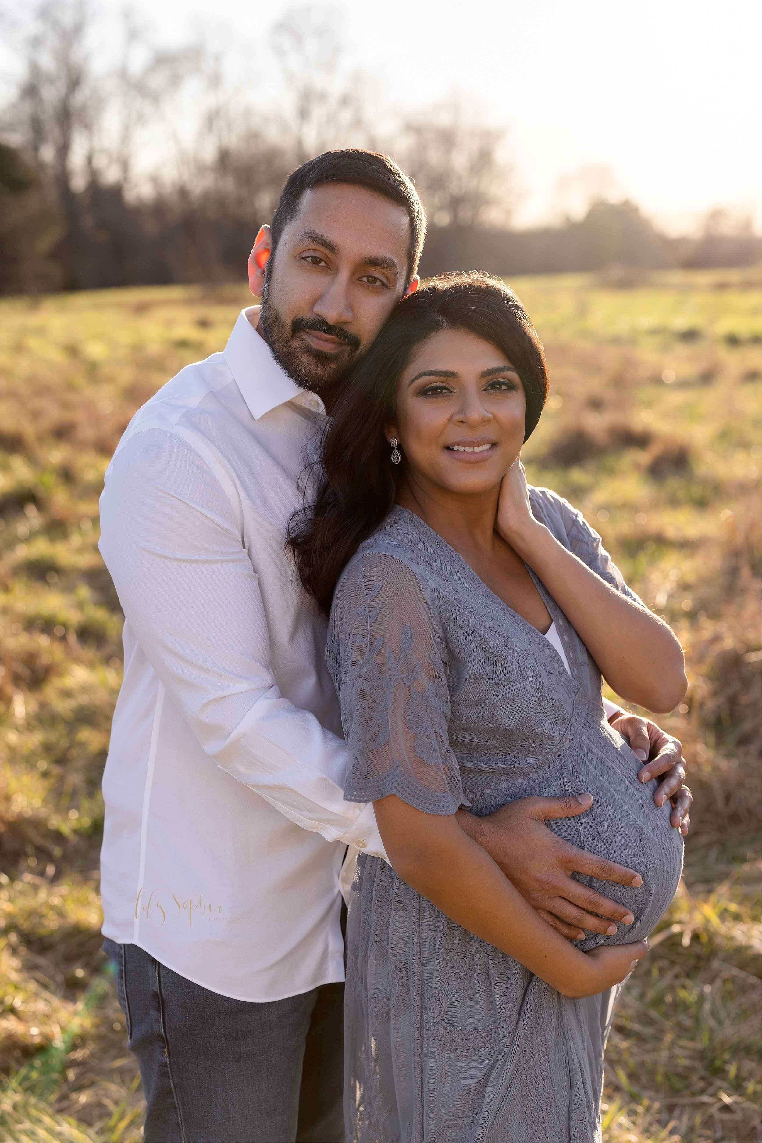  Maternity photograph of a husband standing behind his wife and wrapping his hands around his wife’s waist to place his hands on their child in utero as he rests his head on top of his wife’s head as she holds the base of her belly with her right han