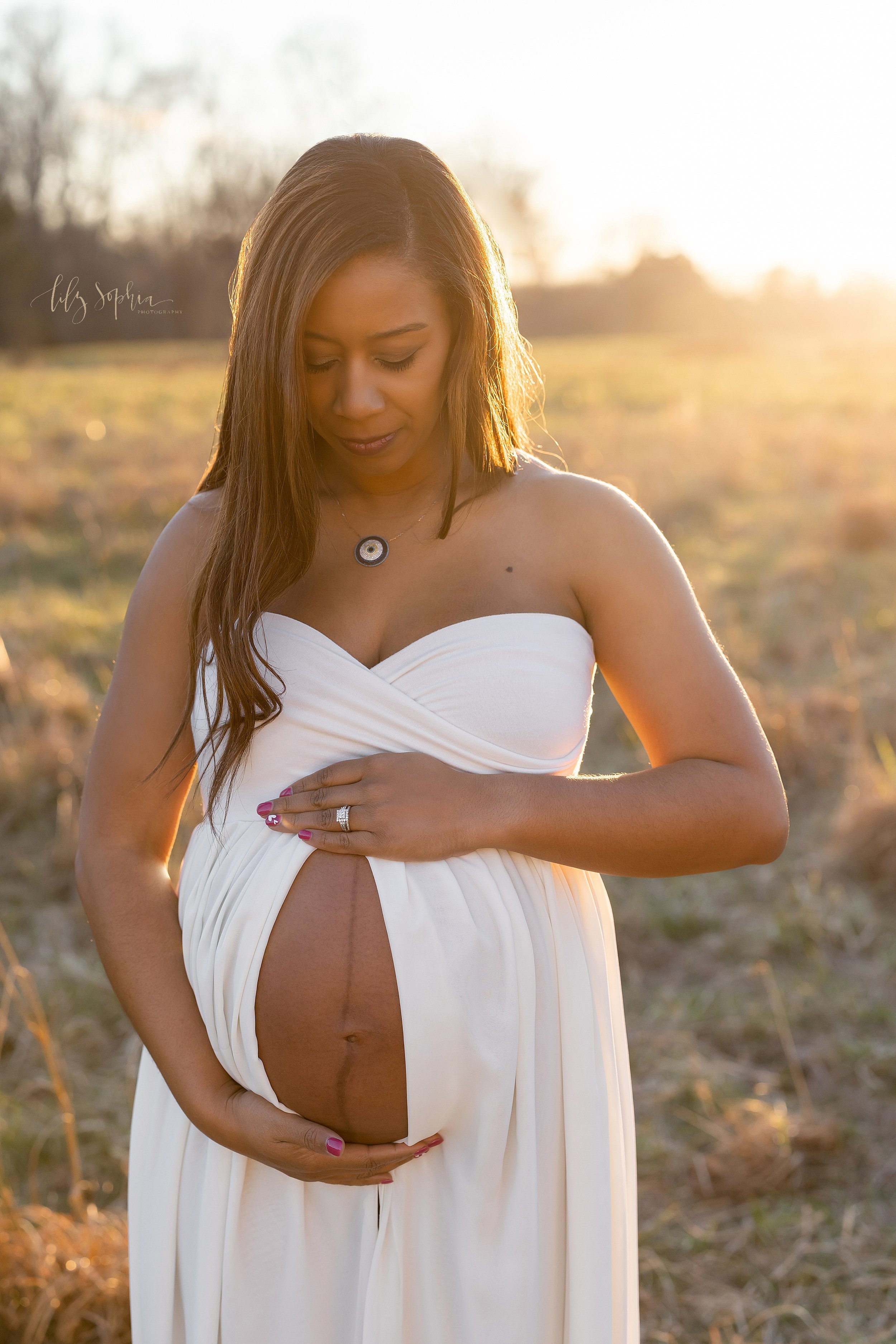  Maternity photograph of a pregnant African-American mother wearing a jersey knit strapless split-front gown holding her left hand on the top of her belly with the dress split open and her right hand at the bass of her belly as she stands in a field 