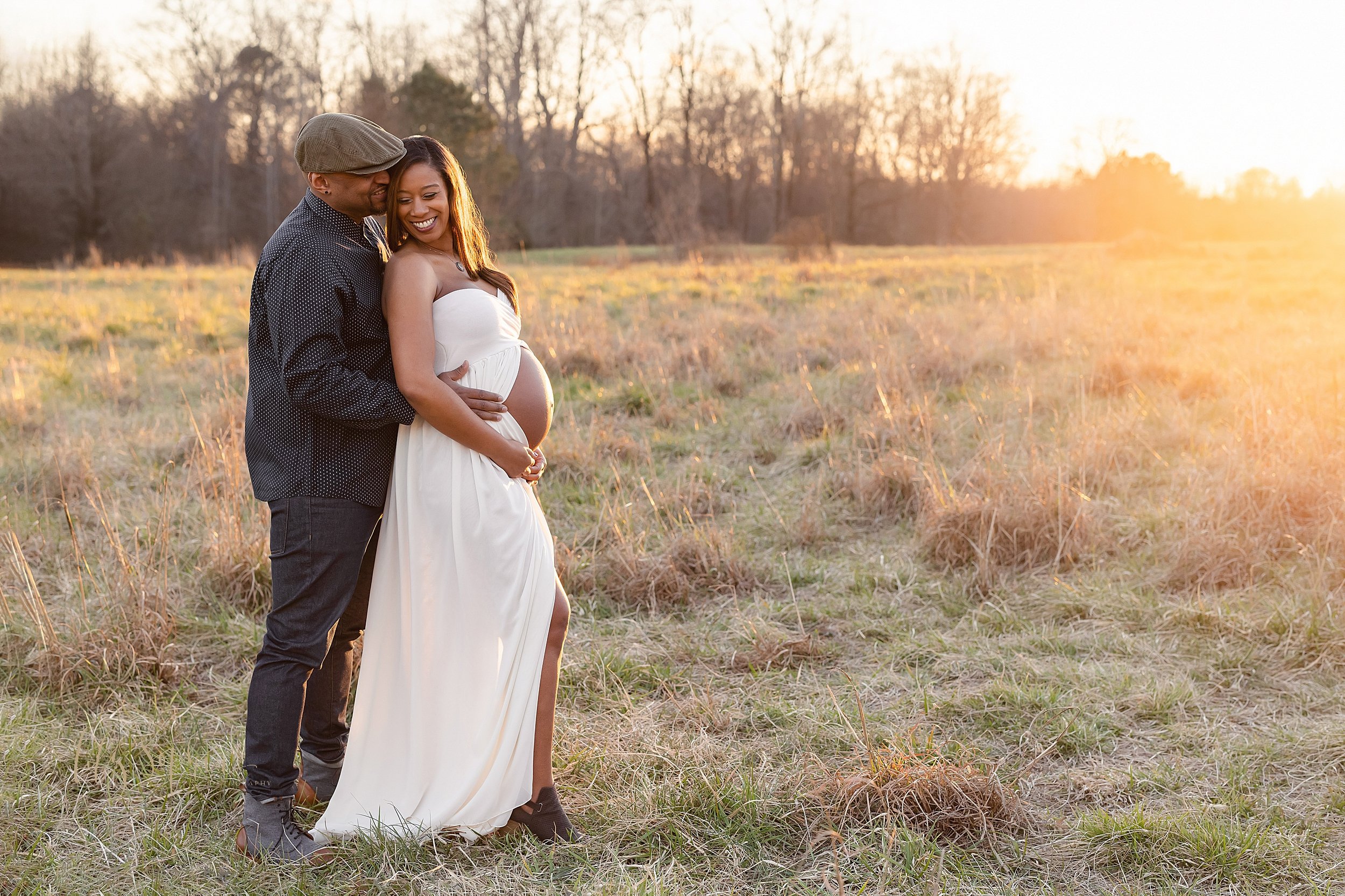  Maternity photograph of an African-American couple standing in a field with the husband behind his wife and his wife opens her strapless jersey knit split-front gown to show her bare belly as she holds the base of it taken near Atlanta at sunset in 