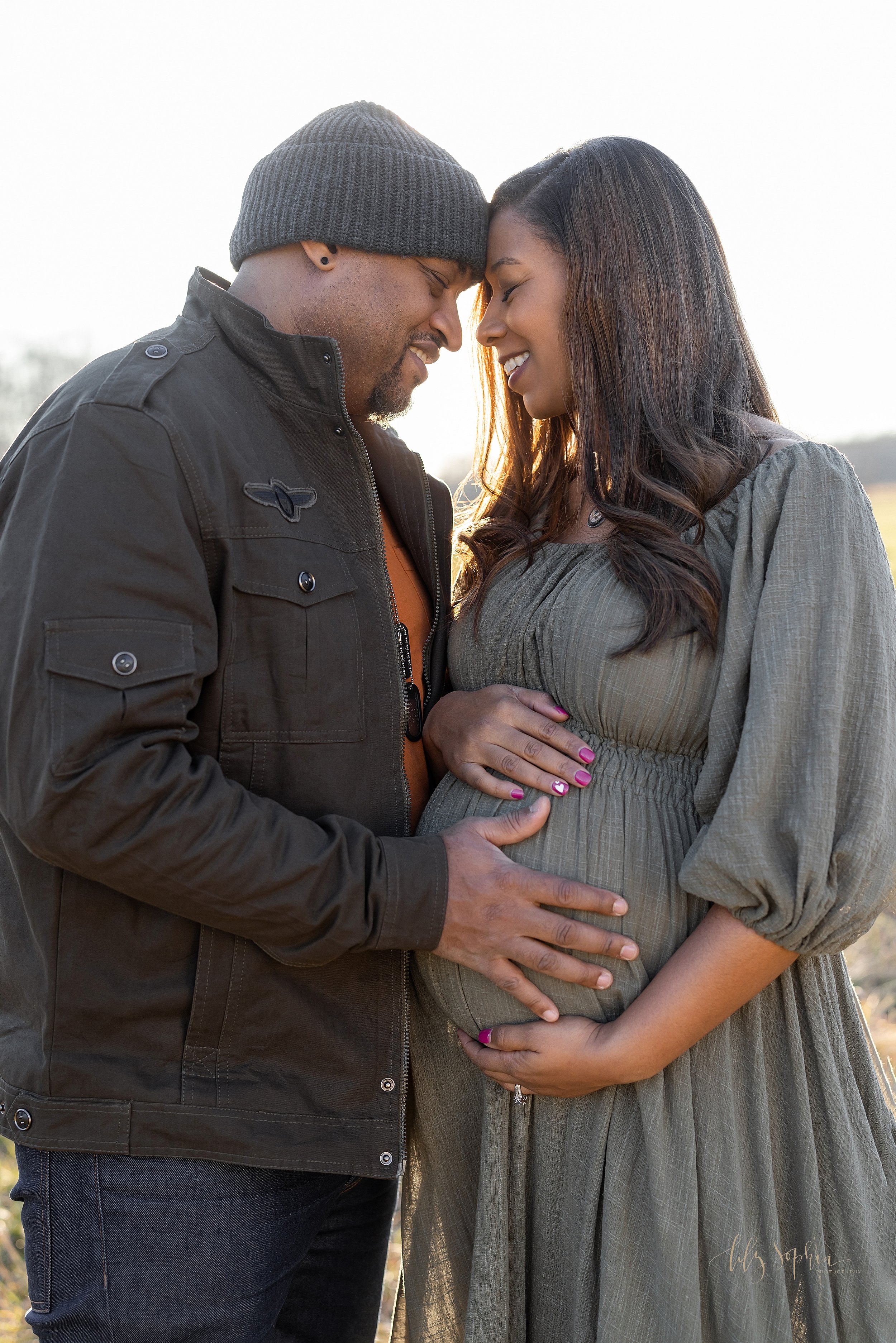  Maternity photo session of an expectant African-American couple during autumn as they stand facing one another with their foreheads together and the father places his hand on his child in utero as the two of them smile in a field near Atlanta at sun