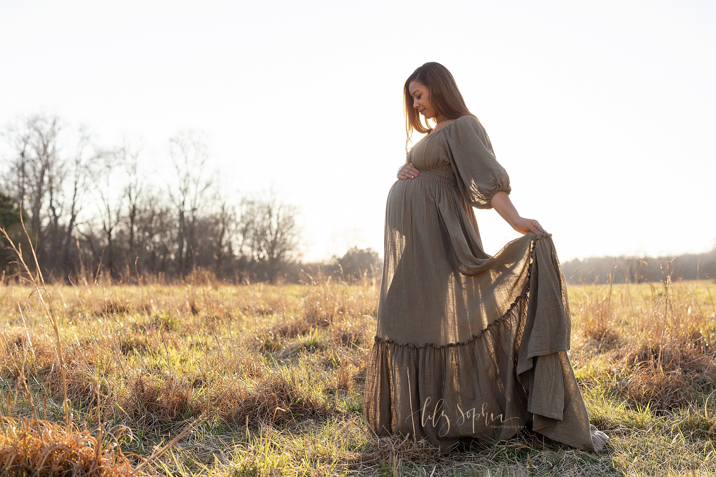  Maternity portrait of a pregnant African-American mother wearing a linen peasant gown as she stands during autumn in an Atlanta field at sunset with her right hand on the top of her belly and her left hand holding the ruffle of the skirt so you can 