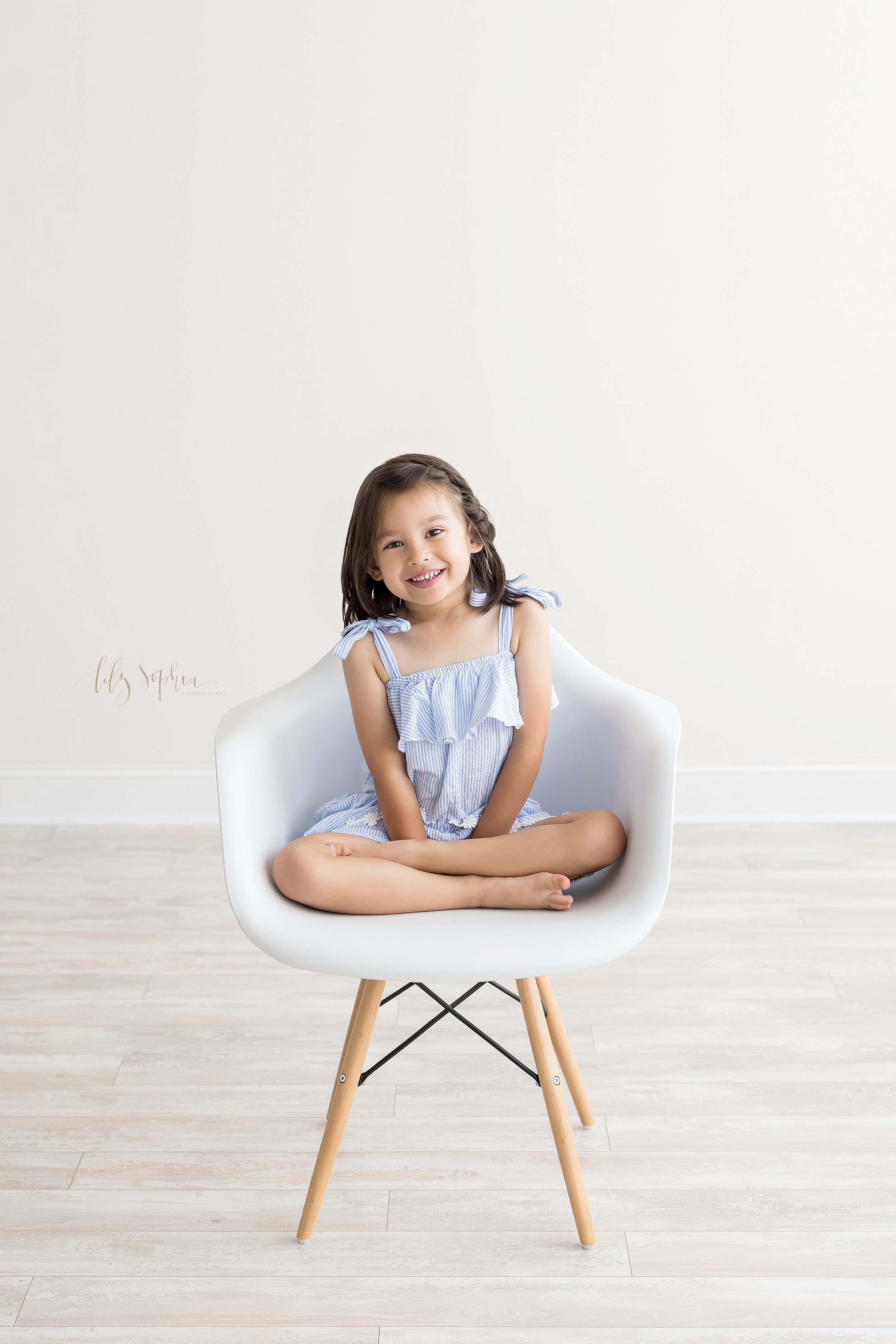  Milestone portrait of a four year old little girl as she sits smiling in a white molded chair taken in a natural light studio near Morningside in Atlanta, Georgia. 