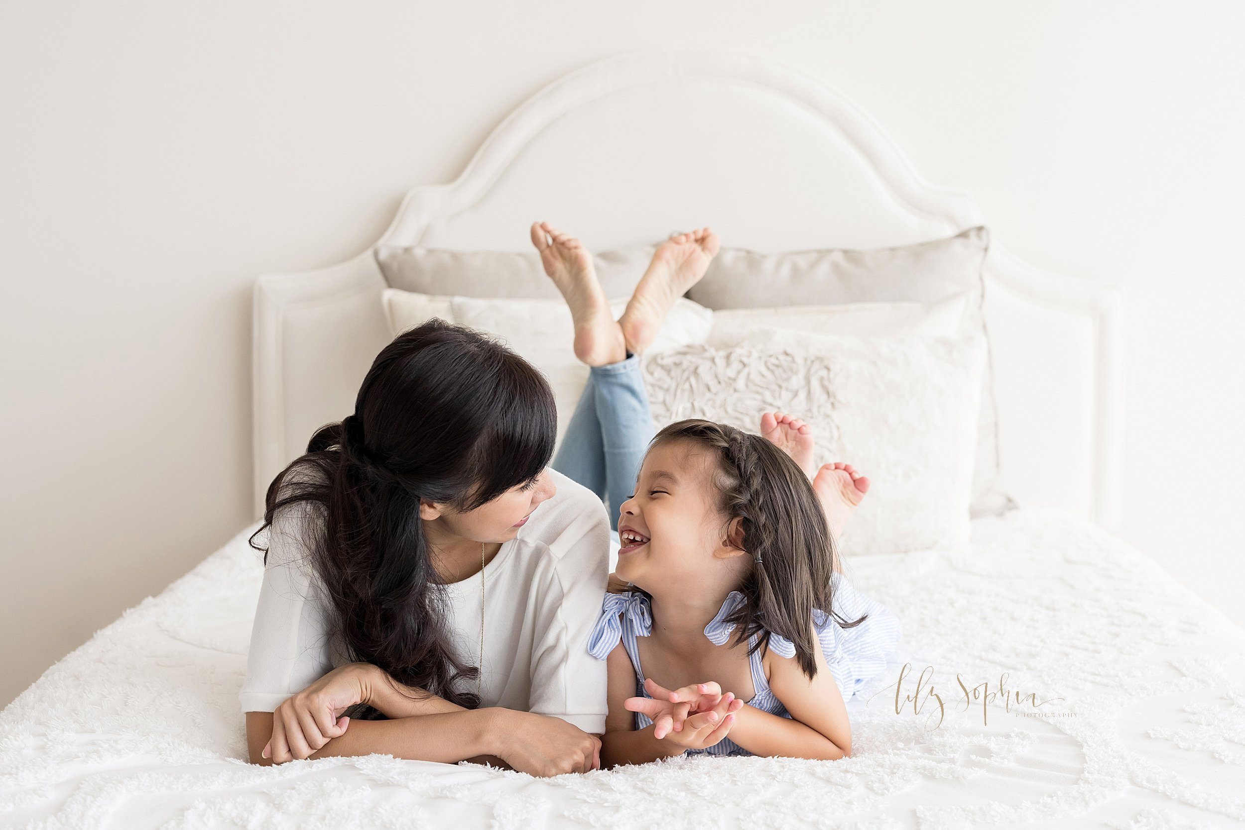  Mother and daughter family photo of an Asian mother and her four year old daughter lying on their stomachs on a bed with their knees bent and their feet in the air as they prop up their bodies with their elbows and chat with one another taken in nat
