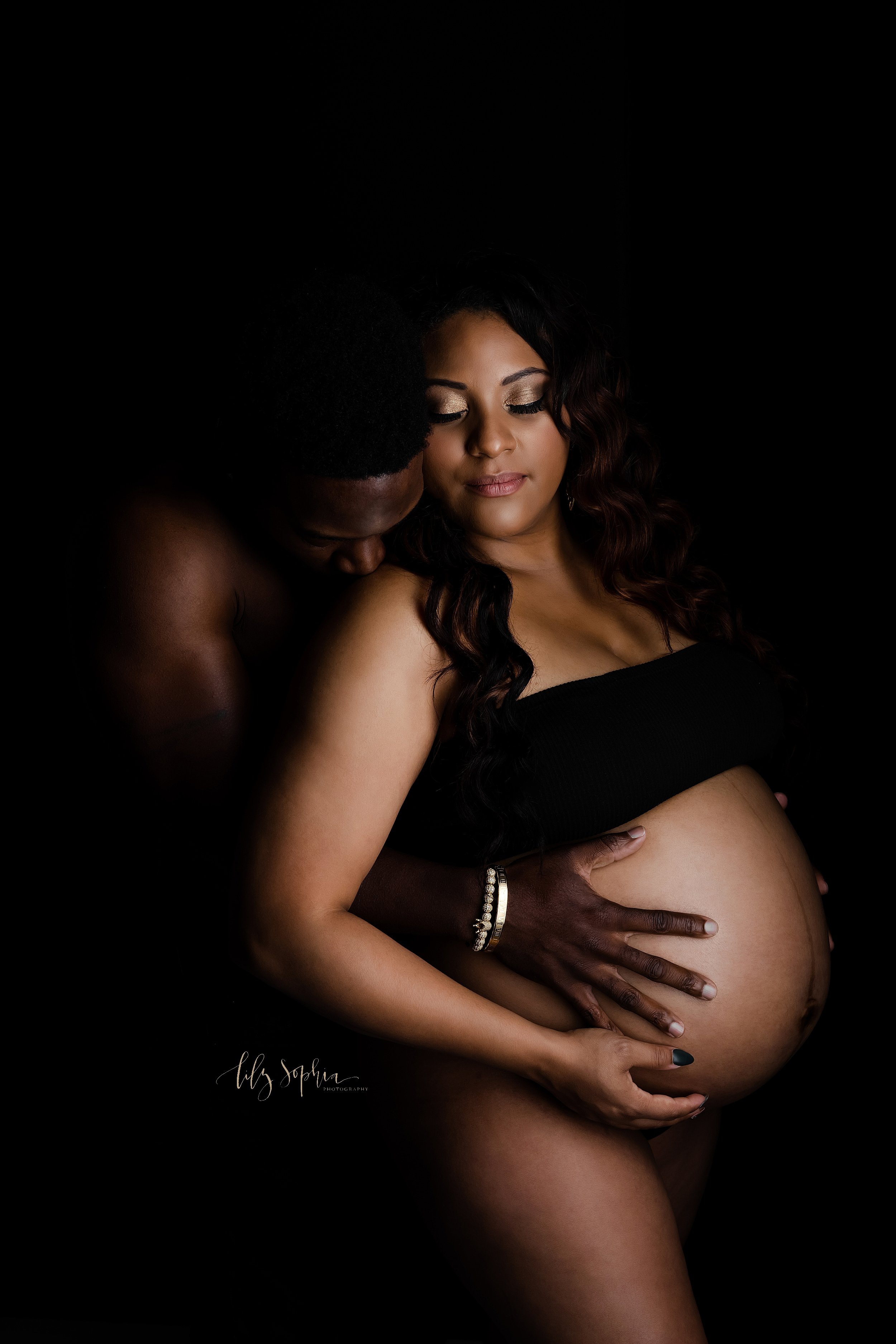  Maternity photograph of an African-American mother as she stands in a studio wearing a black bandeau top and a bare belly as her bare-chested husband stands behind her with his hands on her belly and kisses her on the shoulder taken near Midtown in 