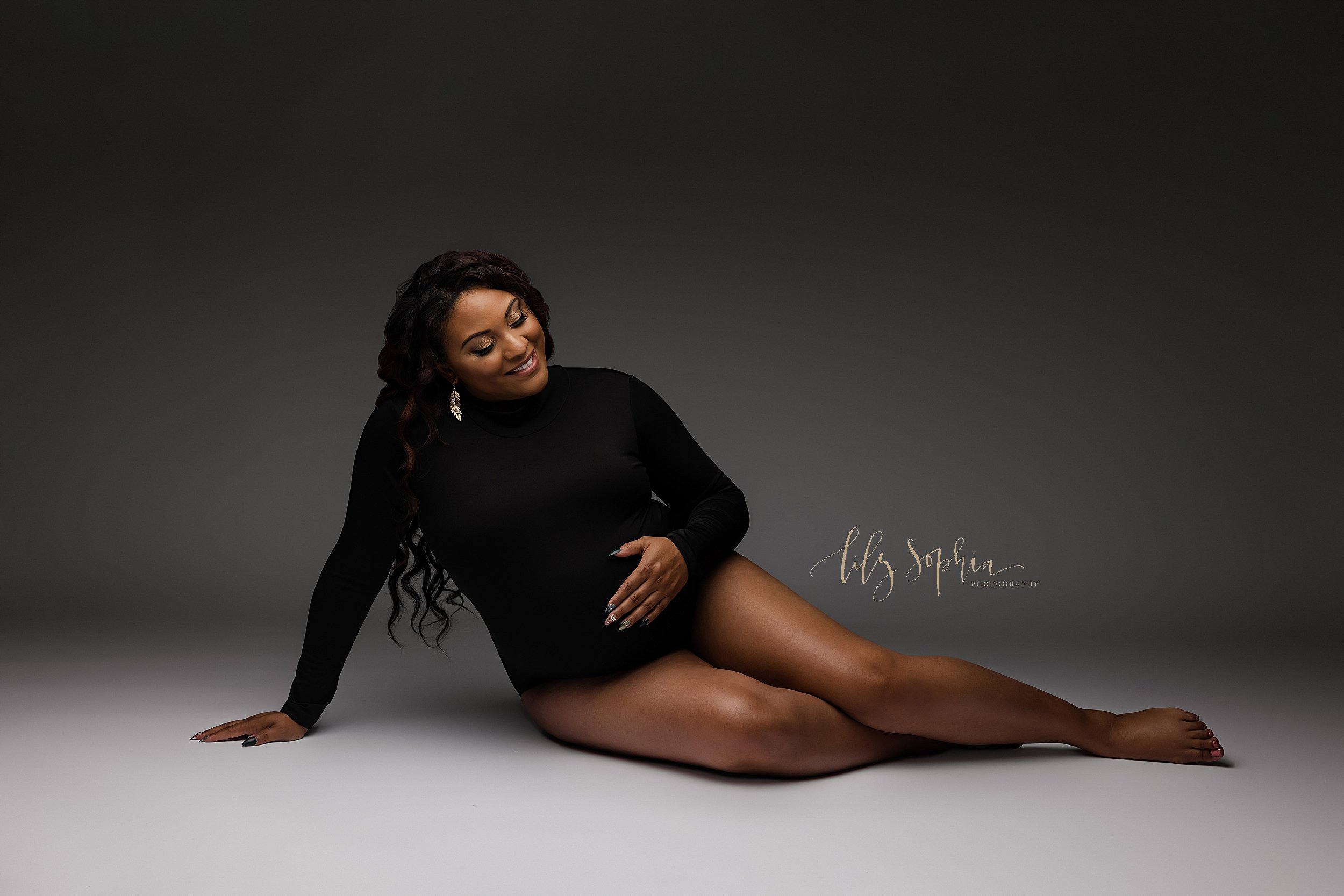  Maternity portrait of a pregnant African-American mother dressed in a black long-sleeved turtle neck sitting on the floor of a natural light studio with her right hand on the floor to hold her up, her left hand on her belly, and her bare legs bent a