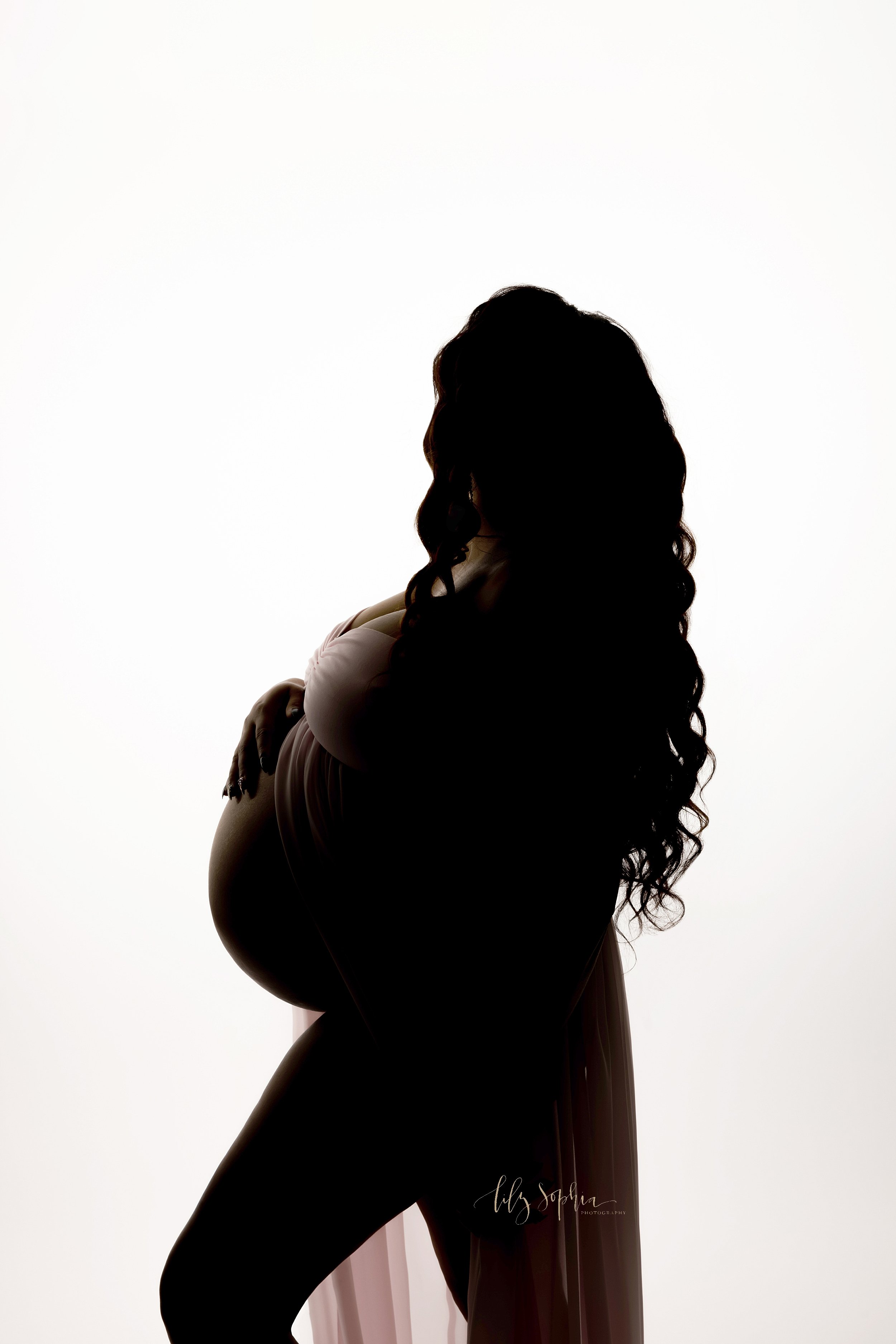  Silhouetted maternity portrait of a pregnant mother as she stands in front of a window streaming natural light wearing a split front gown with her hand on the top of her bare belly as she looks out the studio window taken near Kirkwood in Atlanta. 