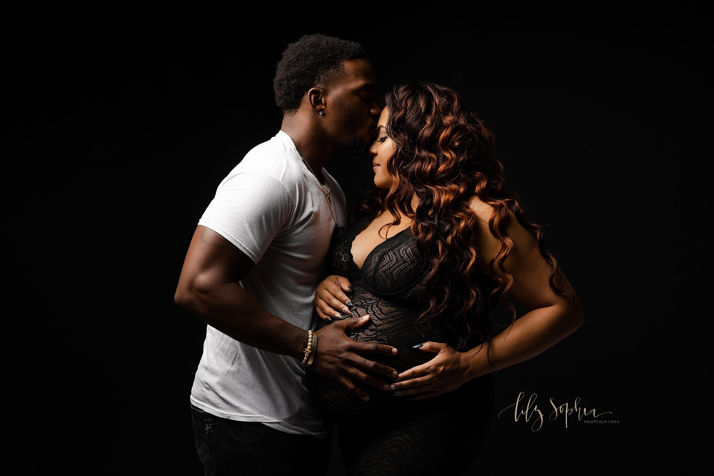  Maternity photo of an African-American mother near Poncey Highlands in Atlanta wearing a black plunging V-neck sheer gown with her  hands on the top and side of her belly as her husband dressed in a white T-shirt faces her, kisses her on the forehea