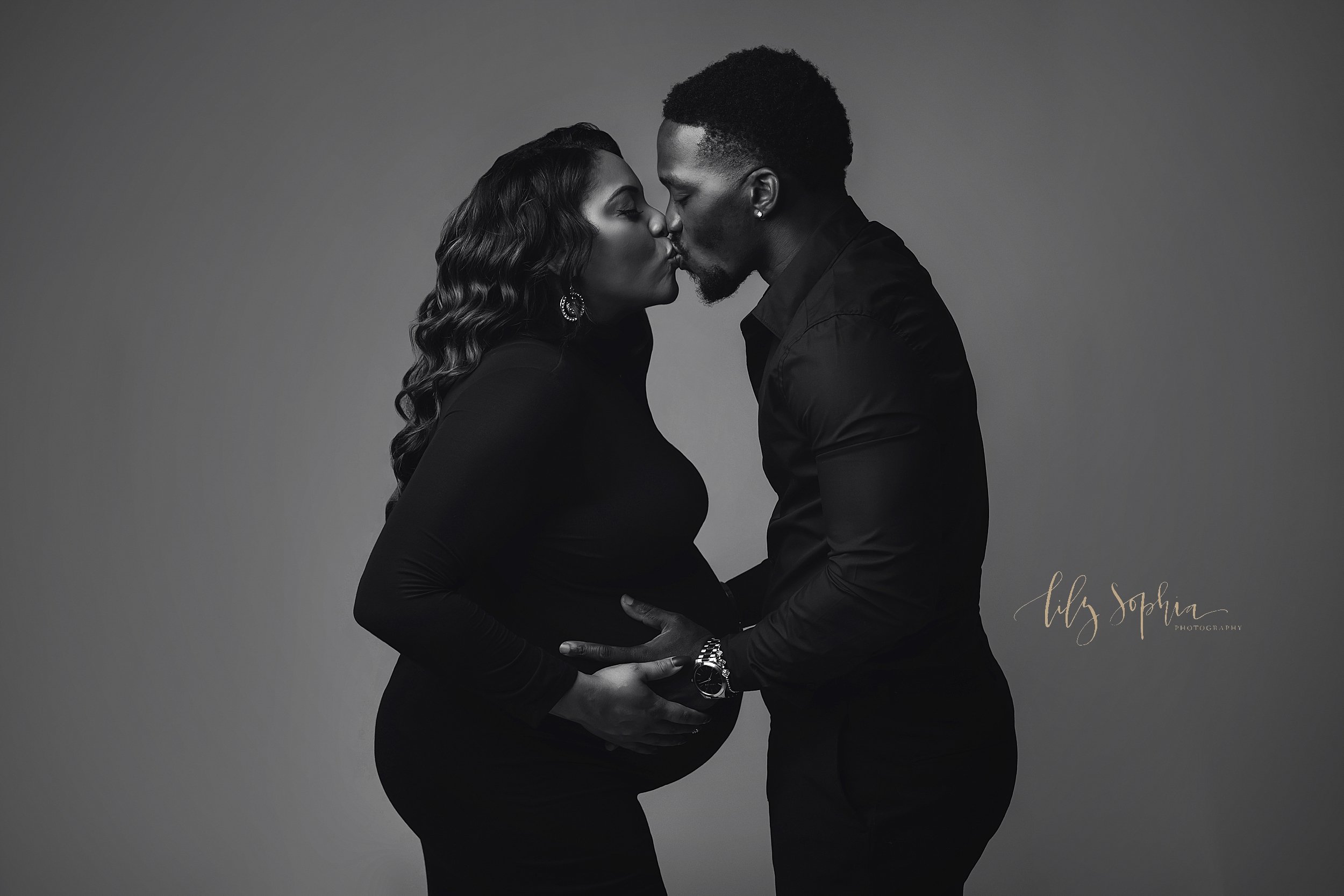  Black and white photograph of an African-American expectant couple dressed in all black as the wife faces her husband and he places his hands to hold their child in utero and she places her hands on top of his while they kiss one another taken near 