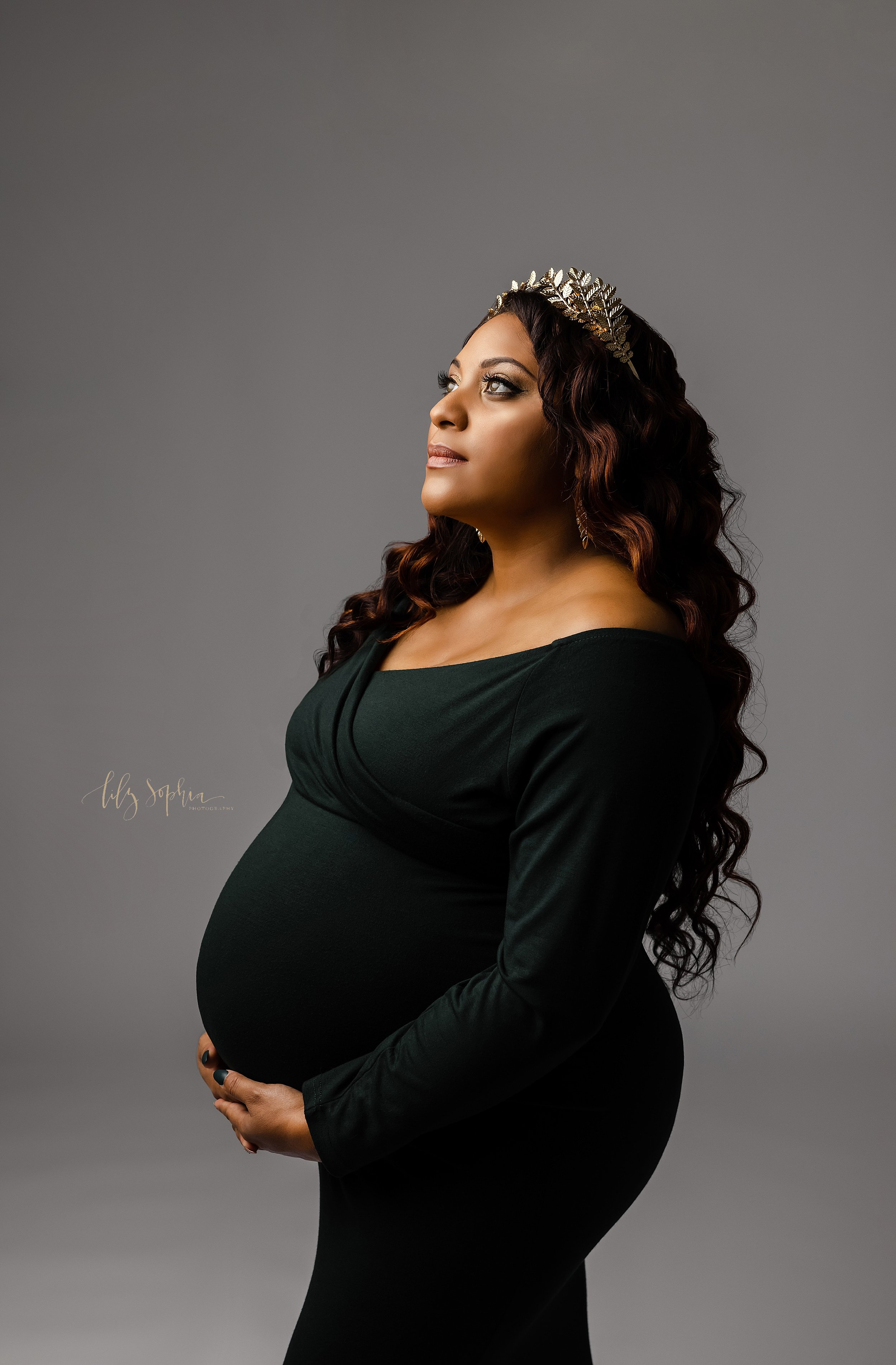  Maternity portrait of an African-American mother wearing a jersey knit long-sleeved black gown and a silver tiara in her flowing curly hair as she stands in a natural light studio near Old Fourth Ward in Atlanta with her hands holding her belly and 