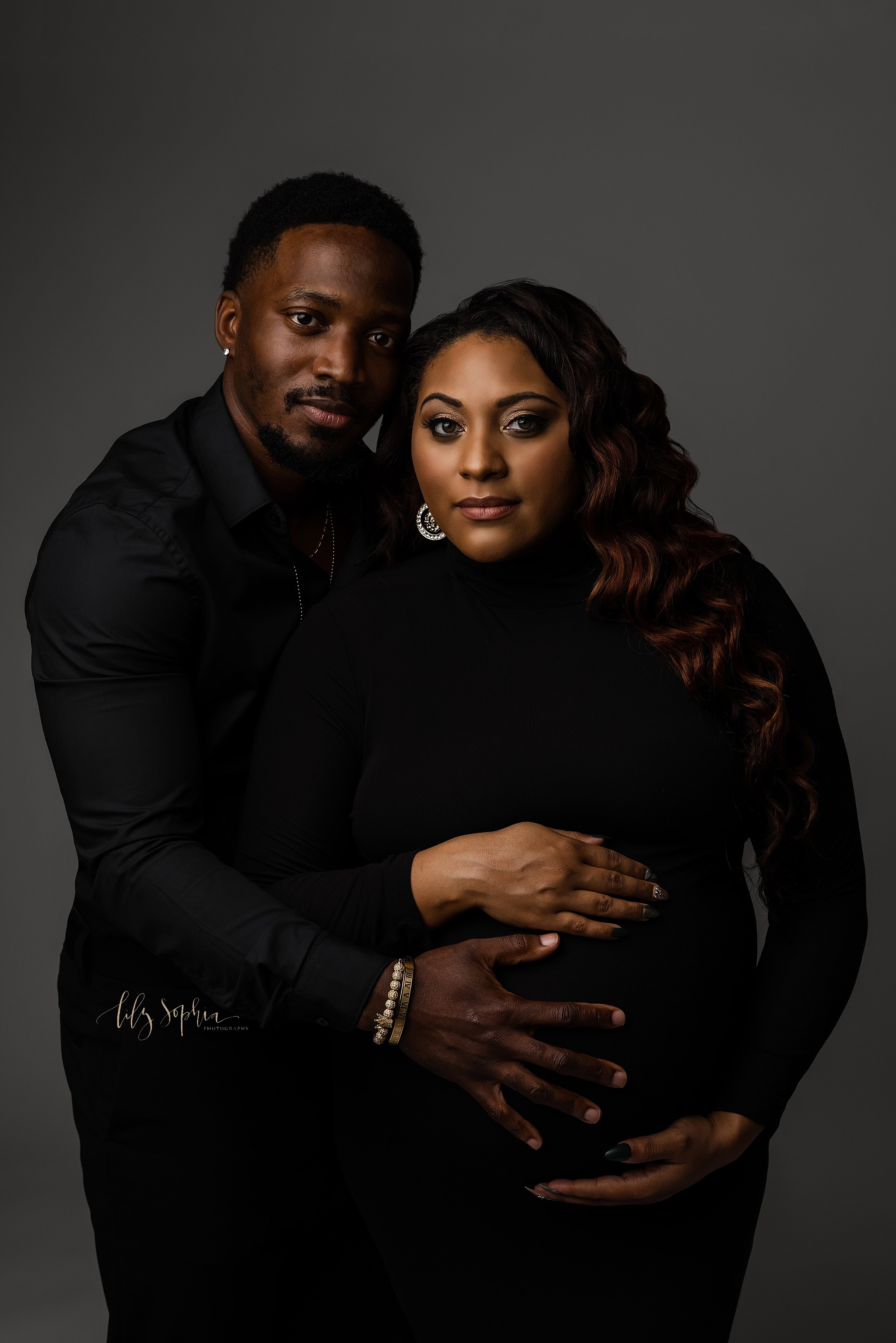  Maternity portrait of an African-American mother dressed in black framing her belly with her hands as her husband stands to her right side and places the palm of his hand on their child in utero taken in a natural light studio in Ponce City Market i