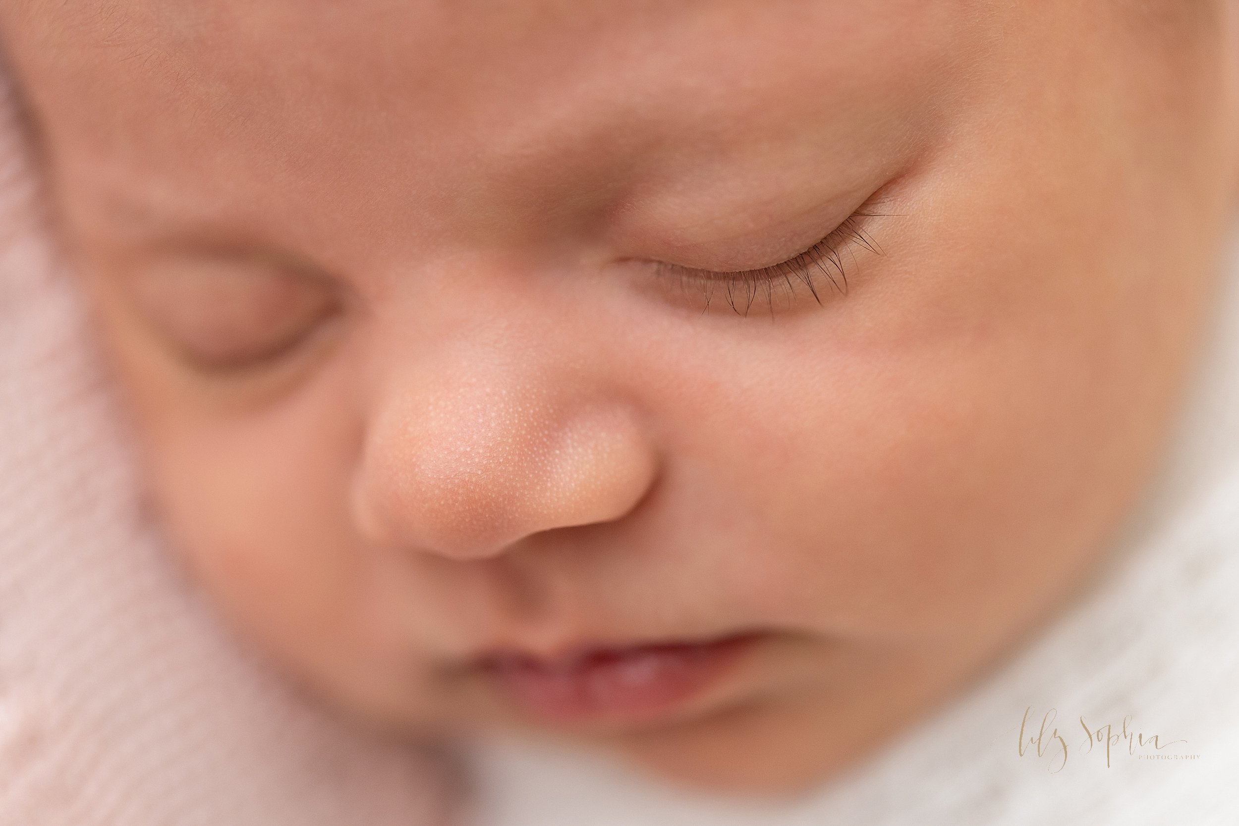  Close-up newborn portrait of the face of a peacefully sleeping baby girl that highlights her button nose, wispy eyelashes, and milky lips taken using natural light near Vinings, Georgia in a studio. 