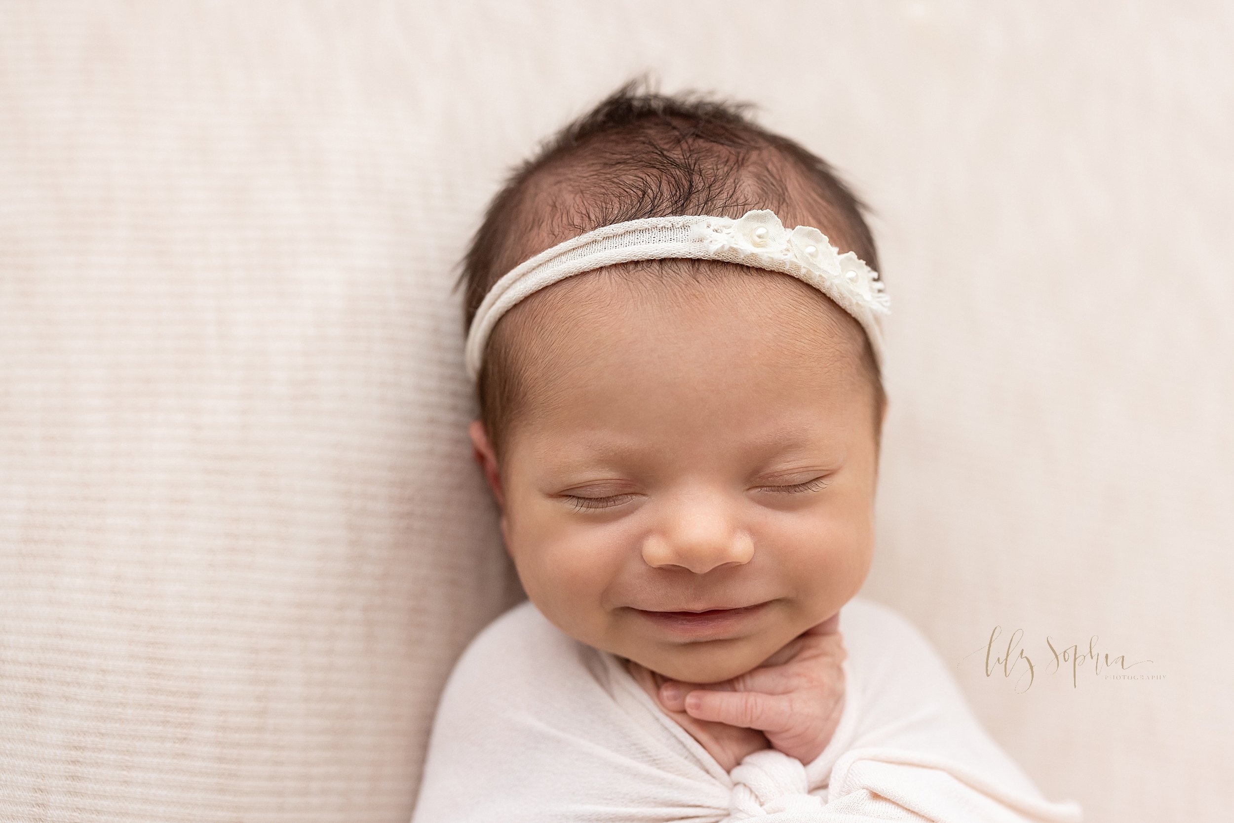  Newborn portrait of an infant girl lying on her back cradled in a stretchy swaddle wearing a jersey headband in her hair with her left hand under her chin as she grins in her sleep taken in a studio near Smyrna, Georgia using natural lighting. 