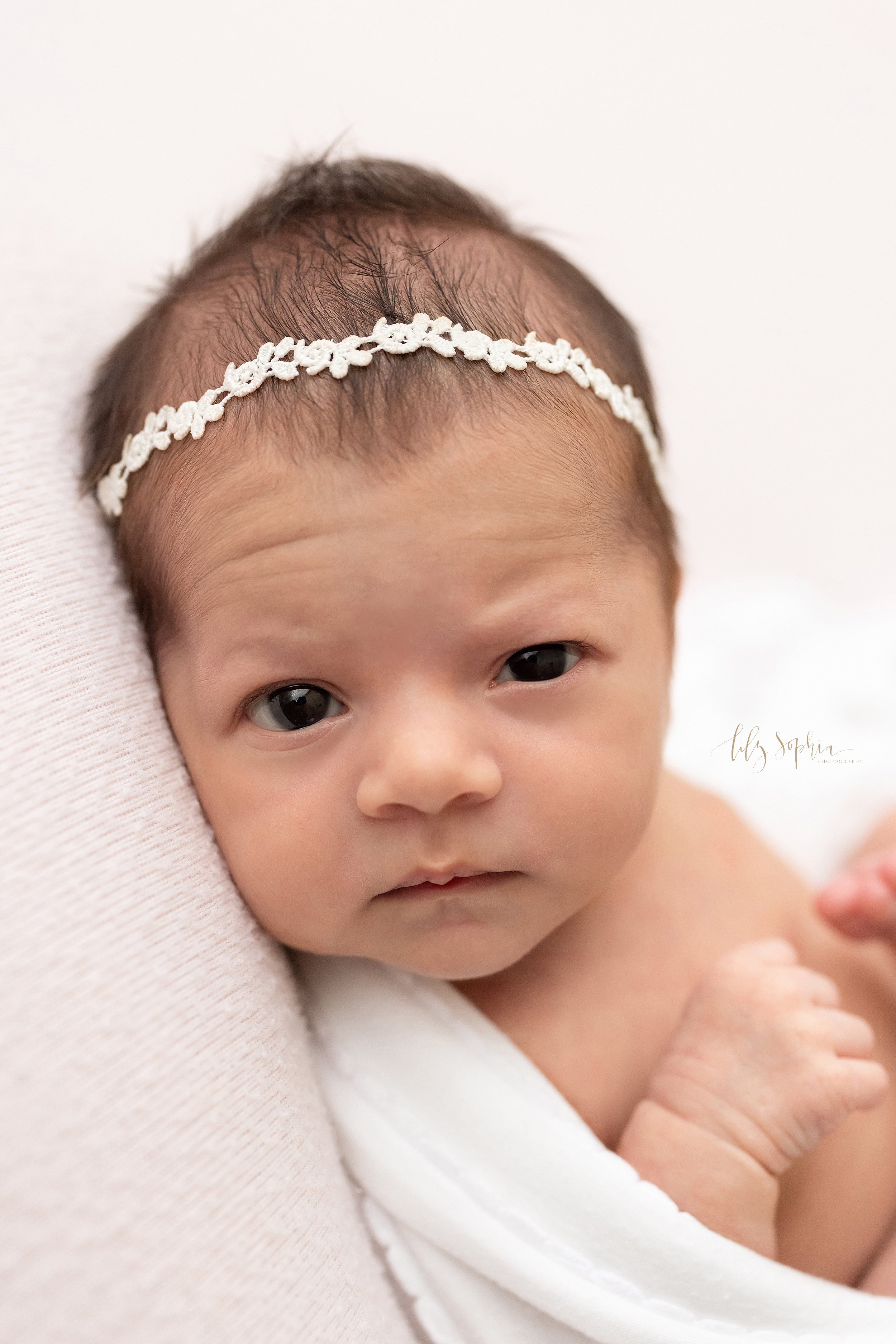  Newborn portrait of a wide awake baby girl wearing a delicate laurel headband in her wispy hair as she lies on her back cradled in a stretchy swaddle taken near Virginia Highlands in Atlanta in a studio that uses natural light. 