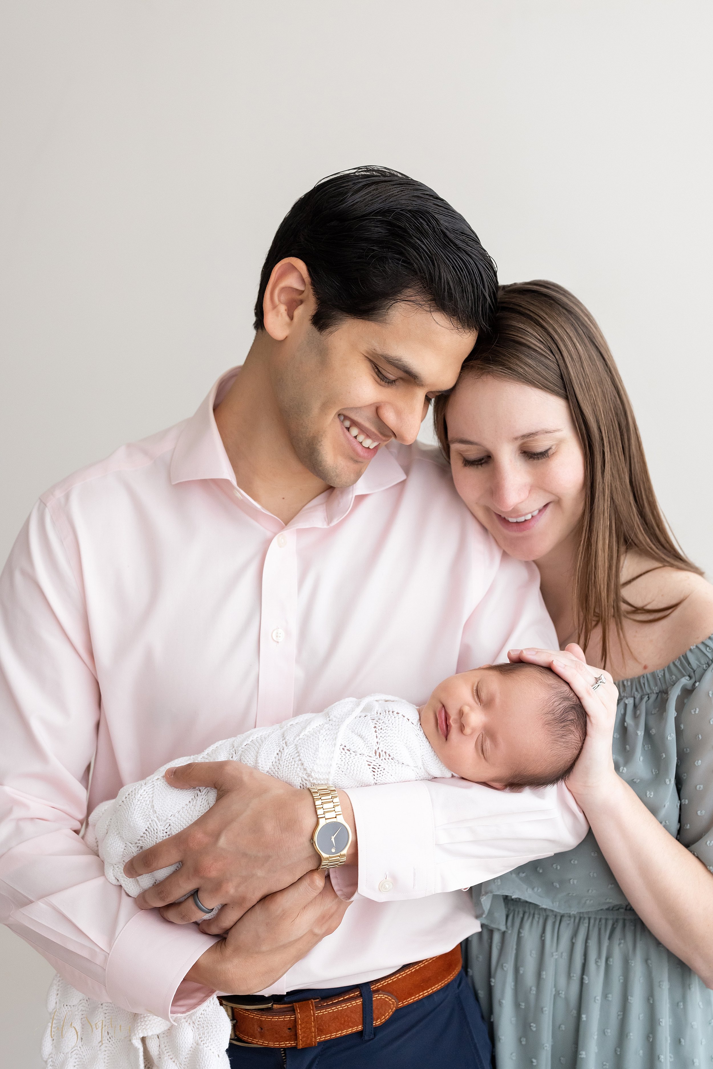  Newborn portrait shoot of a father holding his infant daughter in his arms with his wife standing behind his left shoulder to peer at their child as she places her hand on her daughter’s head taken in a studio using natural light near Alpharetta in 